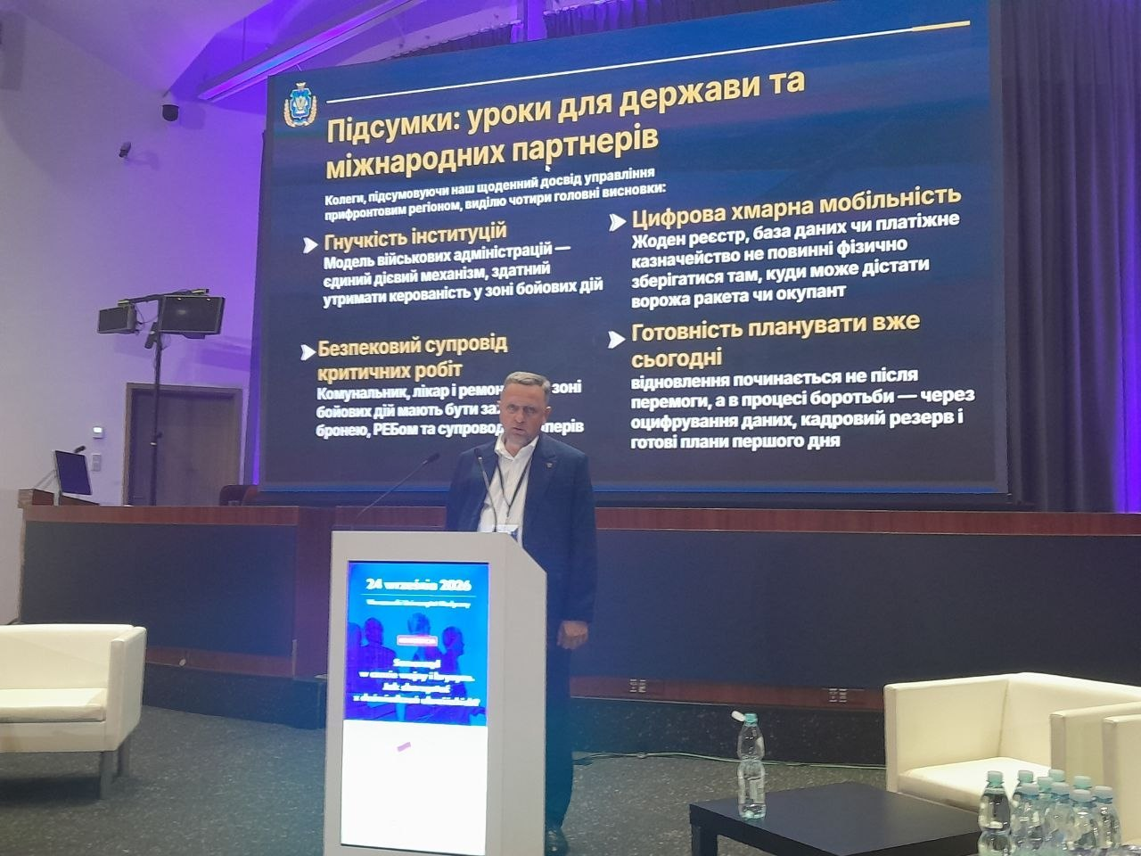 Перший заступник начальника Херсонської ОВА Дмитро Бутрій у класичному темно-синьому костюмі виступає за трибуною на сцені у Варшаві, презентуючи на великому мультимедійному екрані підсумкові уроки кризового адміністрування прифронтових регіонів для міжнародних партнерів. На головному моніторі позаду посадовця відображено презентацію українською мовою з логотипом ХОВА та чотирма ключовими висновками: «Гнучкість інституцій», «Безпековий супровід критичних робіт» (броня, РЕБ для комунальників і медиків), «Цифрова хмарна мобільність» (збереження баз даних у безпеці) та «Готовність планувати вже сьогодні». Перед трибуною з синім банером конференції та датою 24 вересня 2026 року спікер ділиться досвідом протидії атакам FPV-дронів, розгортання антидронових загороджень, облаштування укриттів і децентралізації інфраструктури для збереження життєдіяльності деокупованих громад.