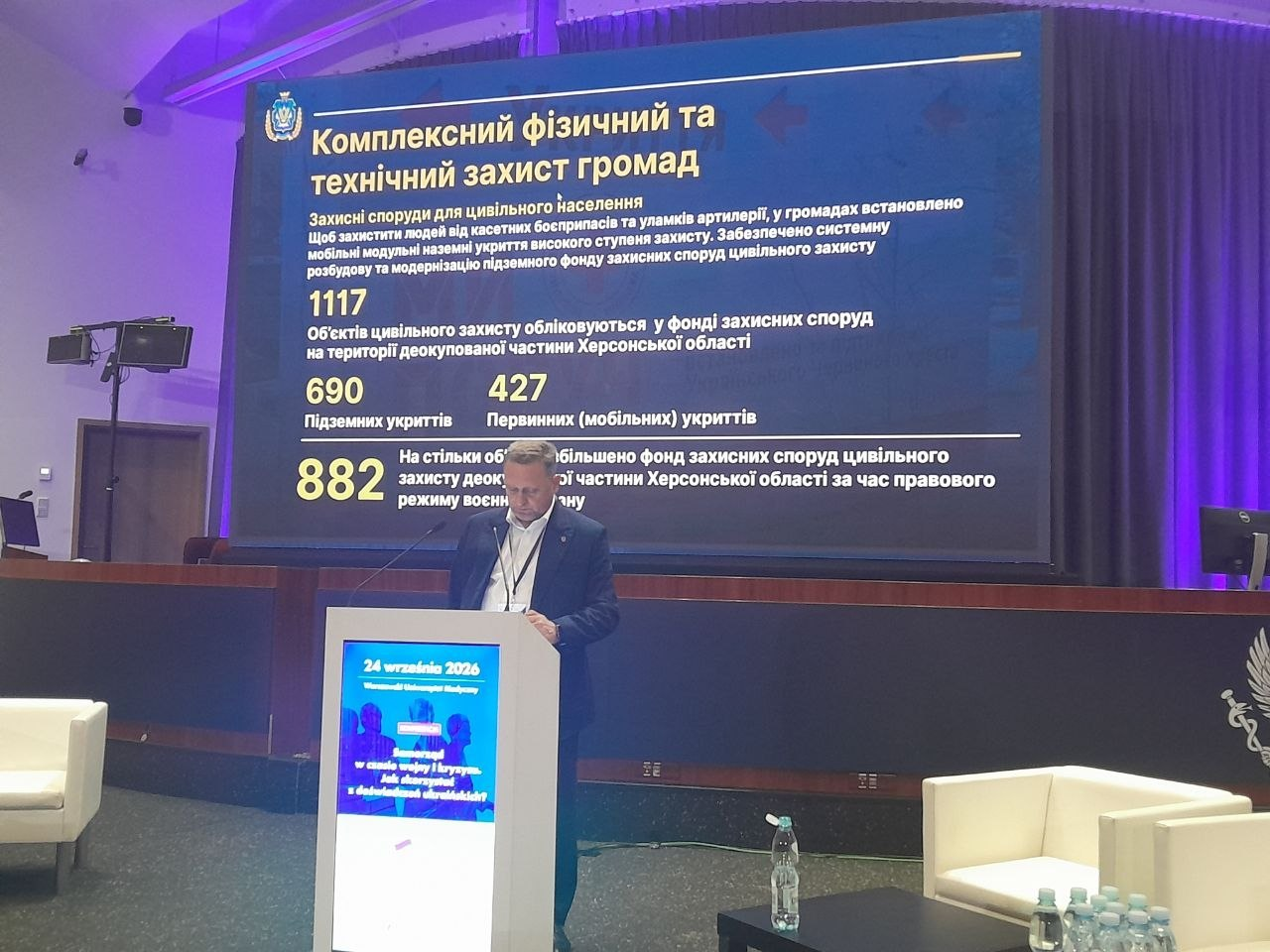 Перший заступник начальника Херсонської ОВА Дмитро Бутрій виступає за трибуною на сцені конференції у Варшаві, презентуючи на великому екрані статистику та досвід розбудови захисних споруд у межах доповіді «Комплексний фізичний та технічний захист громад». На слайді презентації з офіційним гербом ХОВА наведено детальні дані про фонд цивільного захисту на деокупованій частині Херсонщини, що налічує 1117 об'єктів, серед яких 690 підземних та 427 первинних мобільних укриттів, а також зазначено про збільшення цього фонду на 882 об'єкти за час правового режиму воєнного стану. Спікер за кафедрою з синім банером і датою 24 вересня 2026 року ознайомлює польських урядовців та європейських дипломатів із практичними інженерними протоколами захисту населення і критичного обладнання від ударів артилерії, КАБів та системних атак дронів.