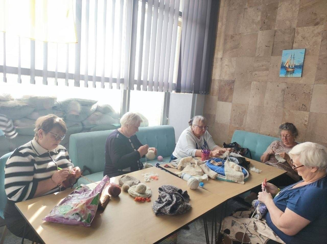 Жінки старшого віку з-поміж переселенок із Херсонщини зосереджено в'яжуть теплі речі за довгим дерев'яним столом у світлому кабінеті в Миколаєві під час занять творчого волонтерського гуртка «Коло тепла». Учасниці зустрічі розмістилися на бірюзових диванах, розклавши перед собою різнокольорові мотки пряжі, спиці, гачки та готові плетені вироби, а стіна праворуч оздоблена каменем та прикрашена картиною з кораблем. Кадр ілюструє щоденну діяльність Координаційного центру «Вільні разом», де літні жінки виготовляють килимки, шкарпетки, маскувальні сітки та окопні свічки для подальшої передачі підрозділам Сил оборони України задля підтримки захисників.