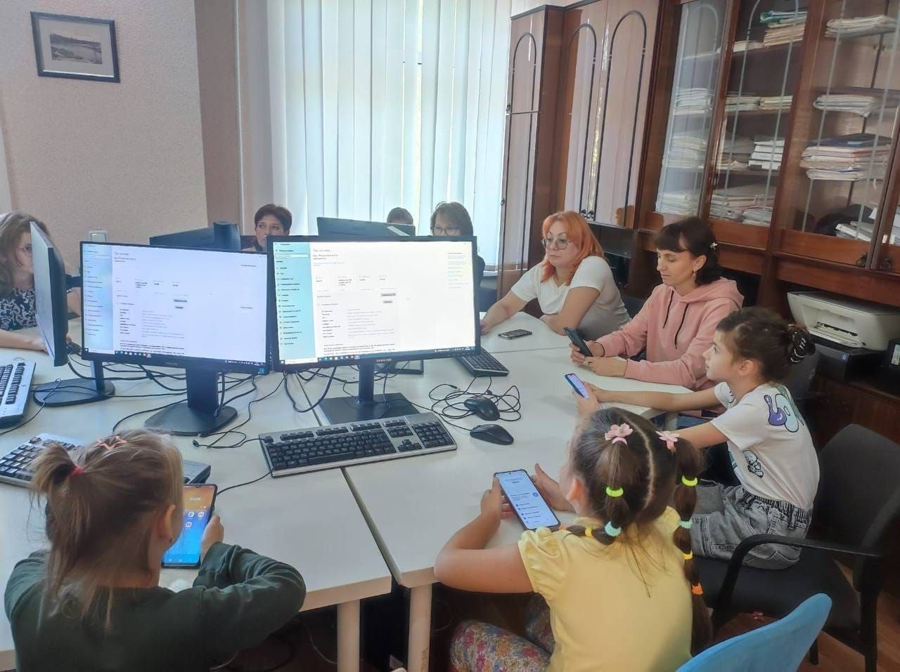 Група жінок та дітей із-поміж переселенців із Херсонщини сидить за великим столом у комп'ютерному класі в Миколаєві під час регулярних занять із комп’ютерної грамотності та вивчення англійської мови, які організував Координаційний центр «Вільні разом». На столі встановлено два великі монітори комп'ютерів із відкритими інтерфейсами навчальних програм, перед якими розкладено клавіатури, а троє дівчаток на передньому плані тримають у руках смартфони для виконання інтерактивних завдань. Кадр ілюструє системну діяльність координаційного хабу з надання безкоштовної освітньої підтримки, розвитку цифрових навичок та інтеграції дорослих і дітей в евакуації.