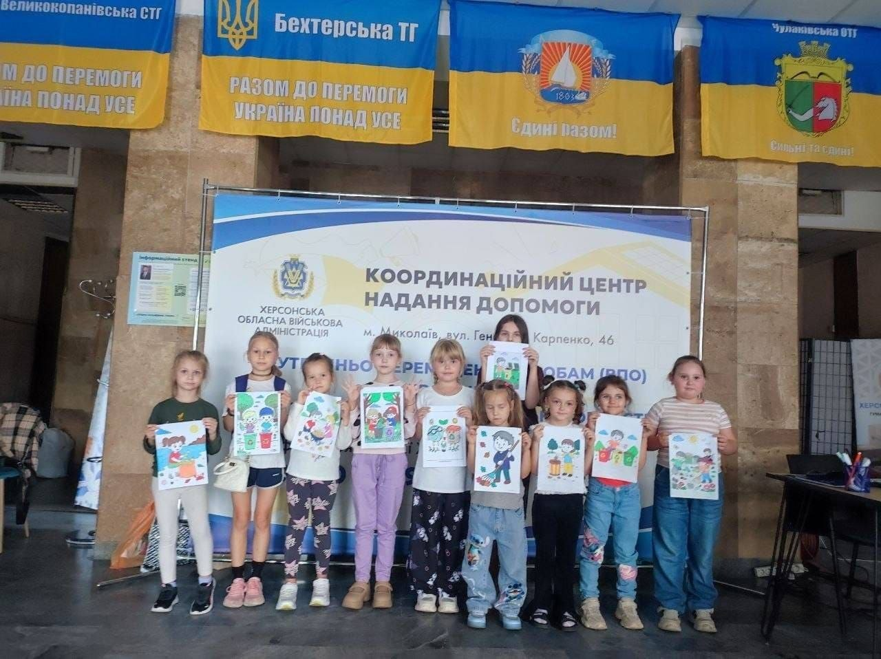 Група дітей стоїть у ряд перед великим банером координаційного центру допомоги в Миколаєві, тримаючи в руках власні малюнки після завершення екологічного конкурсу «Юні захисники довкілля». Діти різного віку посміхаються та позують на тлі великого білого стенда з гербом ХОВА та офіційним написом «КООРДИНАЦІЙНИЙ ЦЕНТР НАДАННЯ ДОПОМОГИ внутрішньо переміщеним особам (ВПО)». У верхній частині кадру над банером розвішано великі Державні Прапори України з геральдикою та назвами прифронтових громад Херсонщини, зокрема Бехтерської ТГ, Великокопанівської СТГ та Чулаківської СТГ, що ілюструє системну діяльність Координаційного центру «Вільні разом» зі збереження зв'язку з рідним краєм, розвитку творчих навичок та екологічної культури малечі в евакуації.