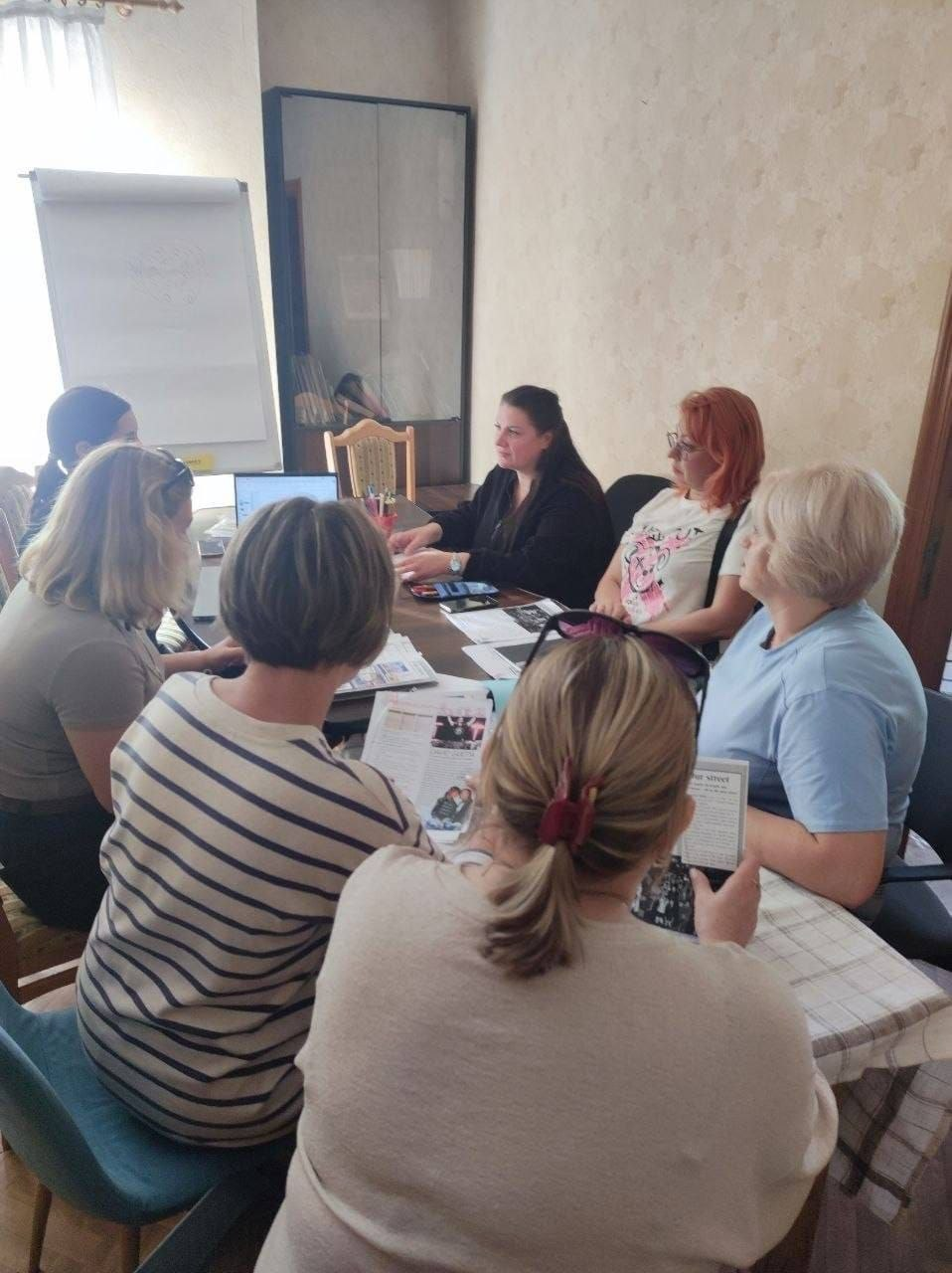 Група жінок із-поміж переселенок із Херсонщини сидить навколо великого столу у кабінеті в Миколаєві під час сесії арттерапії для дорослих, яку організувала команда проєкту «Червона калина» від БО «Українська Освітня Платформа». Учасниці зустрічі вивчають роздруковані інформаційні брошури та матеріали, а модераторка заходу у чорному одязі поруч із відкритим ноутбуком координує обговорення та практичні вправи, спрямовані на психоемоційну підтримку. Засідання проходить у затишній кімнаті з фліпчартом біля вікна, що ілюструє щоденну діяльність Координаційного центру «Вільні разом» із ментального відновлення, пошуку внутрішнього ресурсу та соціальної адаптації громадян в евакуації.