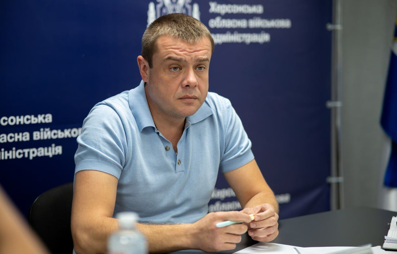Заступник начальника Херсонської ОВА Антон Самойленко у блакитній футболці-поло сидить за столом у залі засідань і тримає в руках ручку під час робочої наради щодо підтримки громад області у сфері комплексного просторового планування. Посадовець зафіксований на тлі фірмового темно-синього пресволу з офіційними написами «Херсонська обласна військова адміністрація», що ілюструє процес координації влади, очільників військових адміністрацій деокупованих населених пунктів та науковців ХДАЕУ, під час якого учасники обговорили залучення потенціалу університету через платформу Living Lab для розроблення планів відбудови деокупованої частини регіону.