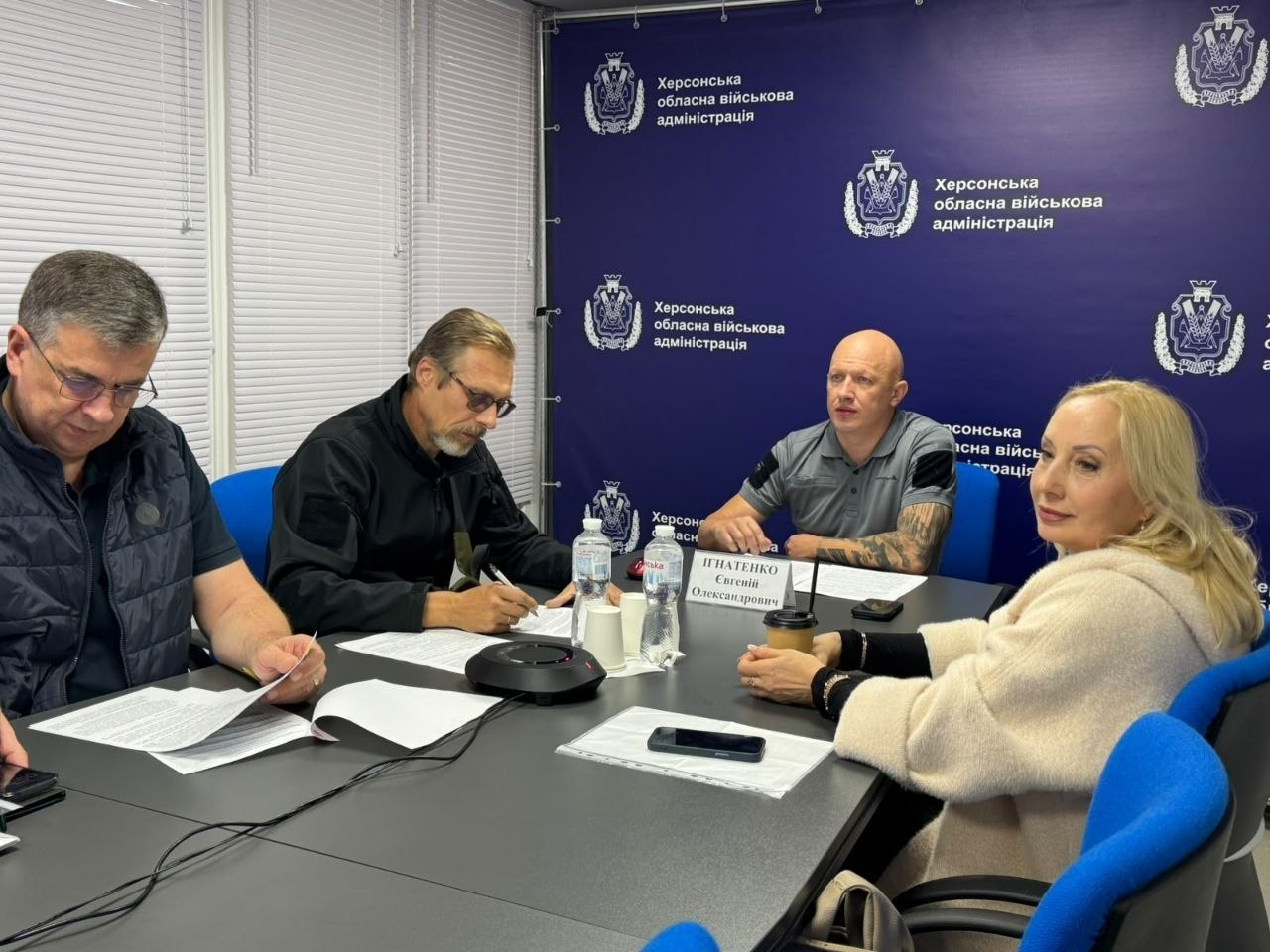 На робочій зустрічі заступник начальника Херсонської ОВА Євгеній Ігнатенко у сірій футболці-поло сидить у центрі столу під час онлайн-засідання Координаційного штабу з проведення евакуаційних заходів та ефективного реагування на масове переміщення населення. По ліву руку від нього розташований директор Департаменту з питань цивільного захисту та оборонної роботи ХОДА Володимир Бєляєв у чорній тактичній кофті, який робить нотатки ручкою, а далі на краю столу сидить перший заступник начальника Херсонської МВА Михайло Линецький в окулярах та темній жилетці, який вивчає роздруковані папери. По праву руку від Ігнатенка, праворуч на передньому плані, сидить директорка Департаменту соціального розвитку Херсонської ОДА Олена Міхєєва у кремовому кардигані з паперовим стаканчиком у руках. Керівники зображені на тлі фірмового темно-синього пресволу з написами «Херсонська обласна військова адміністрація», що ілюструє доповідь під головуванням Міністра у справах громад, територій та внутрішньо переміщених осіб України Віталія Безгіна щодо підготовки транзитного центру в Миколаєві за підтримки УВКБ ООН для можливого посилення евакуації цивільного населення в осінньо-зимовий період.