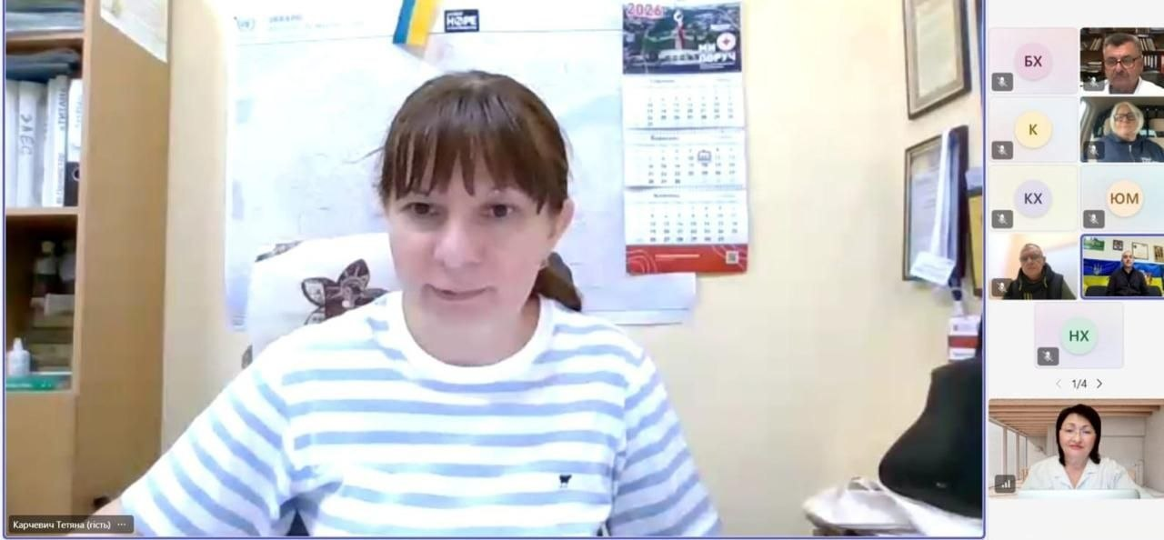 Директорка Департаменту здоров’я Херсонської ОВА Тетяна Карчевич у смугастій біло-блакитній футболці виступає у великому центральному вікні відеодзвінка під час онлайн-засідання Регіональної ради з питань протидії туберкульозу, наркоманії та ВІЛ-інфекції/СНІДу. Посадовець підписана в нижньому лівому кутку як «Карчевич Тетяна (гість)» та зафіксована у робочому кабінеті на тлі настінного календаря на 2026 рік, карти області та маленького прапорця України. У вертикальному правому стовпчику учасників конференції згори донизу відображаються віконця та круглі плашки інших спікерів, серед яких Володимир Рудіч, Наталія Іщенко, представники Чорнобаївської СВА та Галина Коваль у нижньому кутку, що ілюструє координацію між медичними закладами, військовими адміністраціями та громадським сектором для затвердження Регіонального плану відновлення медичних послуг на 2026–2027 роки в умовах воєнного стану.