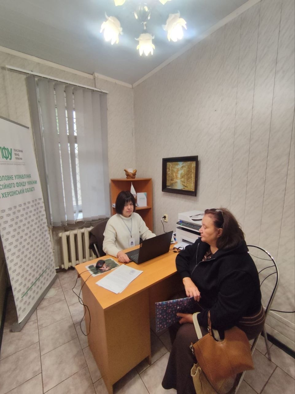Спеціалістка Головного управління Пенсійного фонду України в Херсонській області у світлому светрі за робочим столом консультує відвідувачку з-поміж переселенок із Херсонщини в Одеському координаційному центрі «Вільні разом». Фахівчиня працює за ноутбуком, а літня жінка у чорній кофті сидить навпроти на стільці з папкою та великою сумкою в руках. Ліворуч біля вікна встановлено білий вертикальний ролап із офіційним написом «ГОЛОВНЕ УПРАВЛІННЯ ПЕНСІЙНОГО ФОНДУ УКРАЇНИ В ХЕРСОНСЬКІЙ ОБЛАСТІ», що ілюструє щоденну діяльність хабу та БФ «Сапорт Херсон» із надання виїзних послуг, пенсійного забезпечення та всебічної соціально-правової підтримки херсонців в евакуації.