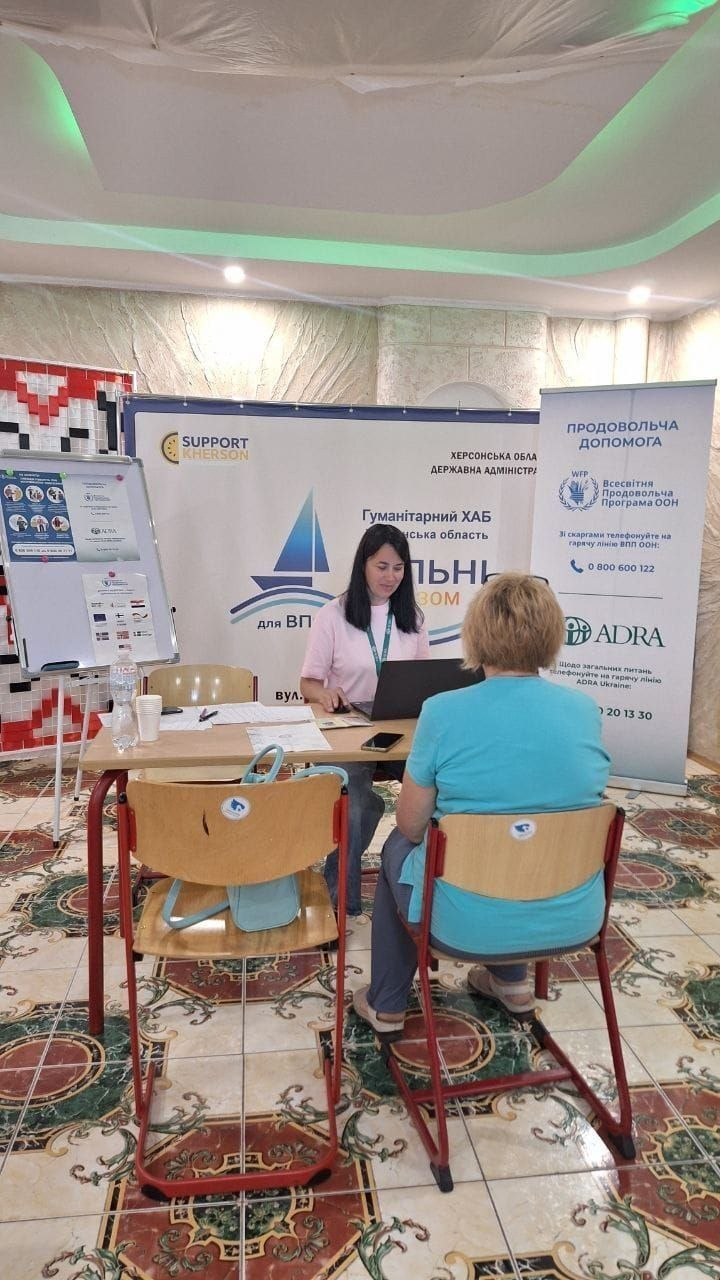 Фахівчиня за робочим столом оформлює документи та реєструє на отримання грошової підтримки літню жінку у блакитній футболці, яка сидить навпроти на стільці спиною до камери. Зустріч проходить у залі Гуманітарного хабу для ВПО «Вільні разом» на тлі великого офіційного банера з логотипами БФ «Support Kherson» та Херсонської ОДА. Праворуч від столу встановлено вертикальний ролап із заголовком «ПРОДОВОЛЬЧА ДОПОМОГА», логотипами Всесвітньої продовольчої програми ООН (ЗПП ООН / WFP) та благодійної організації ADRA, а ліворуч стоїть мобільна дошка-фліпчарт з інформаційними плакатами. Кадр ілюструє спільну діяльність Одеського координаційного центру та благодійників із системної соціально-економічної підтримки новоприбулих переселенців із Херсонщини в евакуації.