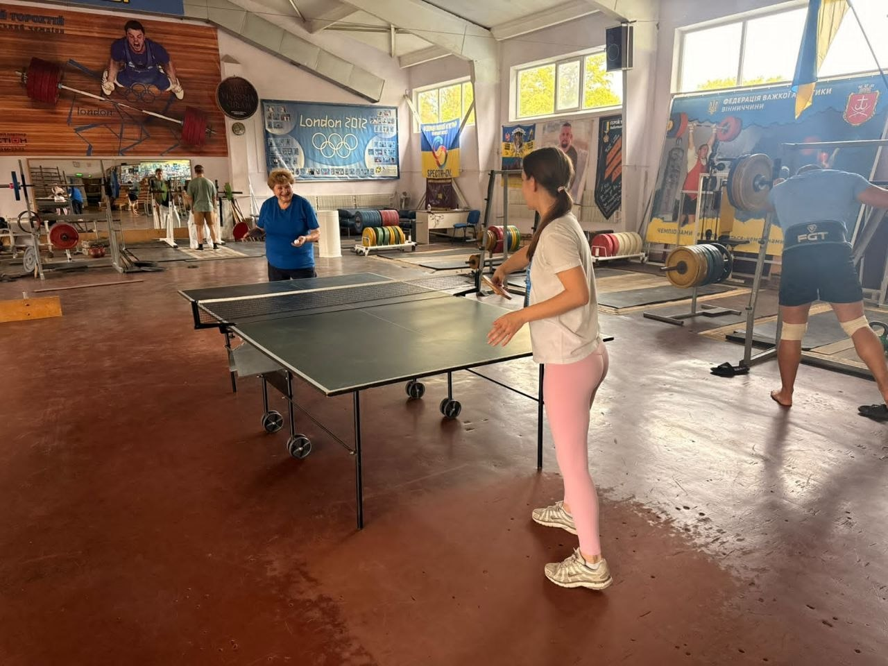 Дві жінки різного віку захоплено грають у настільний теніс по центру просторого спортивного залу у Вінниці під час чергового дружнього тренування для любителів активного відпочинку з-поміж переселенців із Херсонщини. На передньому плані праворуч стоїть молода дівчина у білій футболці та рожевих лосинах із тенісною ракеткою в руці, яка готується відбити м'яч, а навпроти неї за зеленим тенісним столом позицію займає усміхнена літня жінка у синій футболці. Подія проходить у тренувальному залі зі штангами, помостами та дисками для важкої атлетики на тлі великих вікон, а стіни прикрашені олімпійським банером «London 2012» та прапором України, що ілюструє системну діяльність Вінницького координаційного центру «Вільні разом» із популяризації здорового способу життя, соціальної адаптації та психоемоційного розвантаження громадян в евакуації.