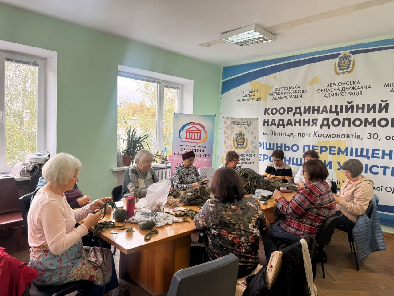 Група жінок різного віку з-поміж переселенок із Херсонщини зосереджено плете захисні нашоломники для українських Захисників та Захисниць, сидячи навколо великого дерев'яного столу в хабі у Вінниці. Учасниці волонтерського процесу розклали перед собою мотки зелених та коричневих ниток, стрічки й заготовки для маскувального спорядження у світлій кімнаті з великими вікнами. Засідання майстринь проходить на тлі великого офіційного банера з гербом ХОДА та написами «КООРДИНАЦІЙНИЙ ЦЕНТР НАДАННЯ ДОПОМОГИ внутрішньо переміщеним особам ХЕРСОНЩИНИ у місті Вінниця», що ілюструє системну діяльність Вінницького координаційного центру «Вільні разом» зі згуртування громади, організації корисного дозвілля та регулярної допомоги Силам оборони України.