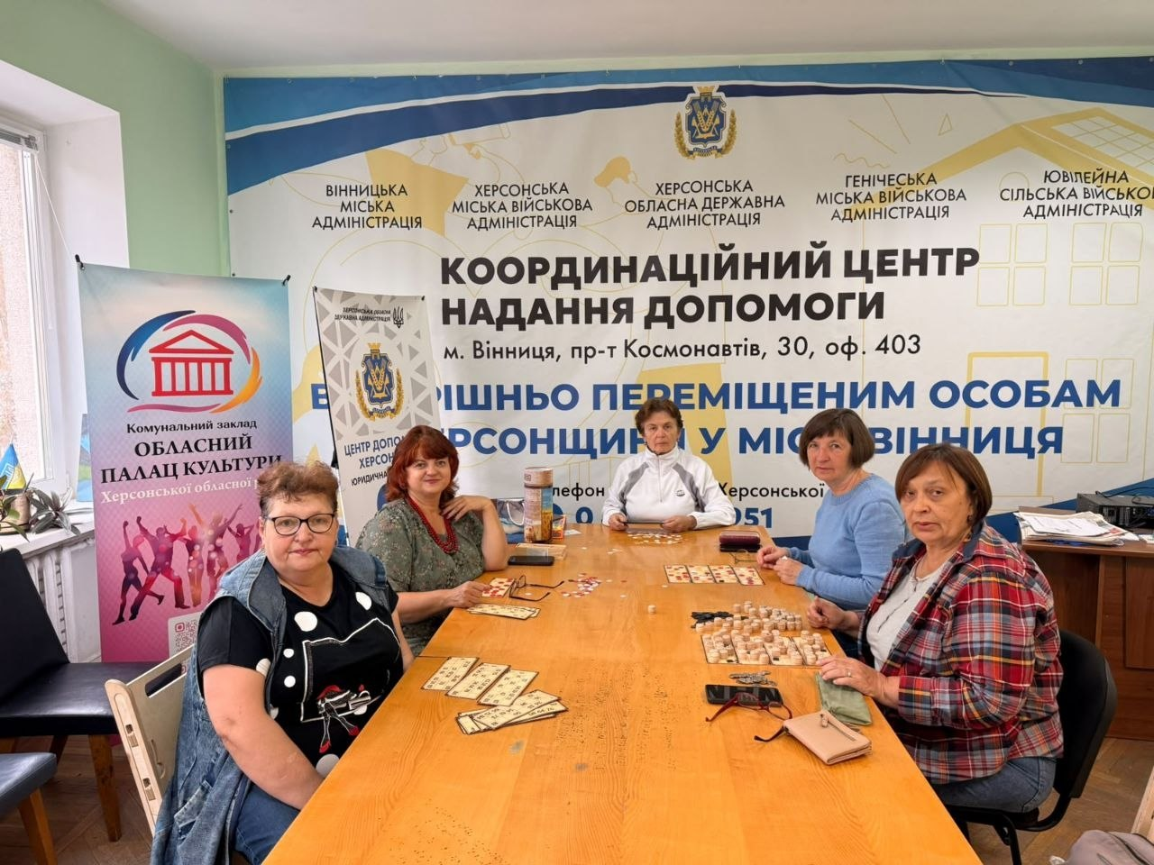 П'ять жінок старшого віку з-поміж переселенок із Херсонщини захоплено грають у настільну гру в лото, сидячи навколо довгого дерев'яного столу в координаційному хабі у Вінниці. Учасниці дозвілля розклали перед собою спеціальні ігровій картки з цифрами та дерев'яні барила у затишній світлій кімнаті біля вікна. Зустріч проходить на тлі великого офіційного банера з гербом ХОДА та написом «КООРДИНАЦІЙНИЙ ЦЕНТР НАДАННЯ ДОПОМОГИ внутрішньо переміщеним особам ХЕРСОНЩИНИ У МІСТІ ВІННИЦЯ», а ліворуч встановлено яскравий ролап обласного Палацу культури, що ілюструє системну діяльність Вінницького координаційного центру «Вільні разом» з організації корисного відпочинку, згуртування земляків та психоемоційного розвантаження громадян в евакуації.