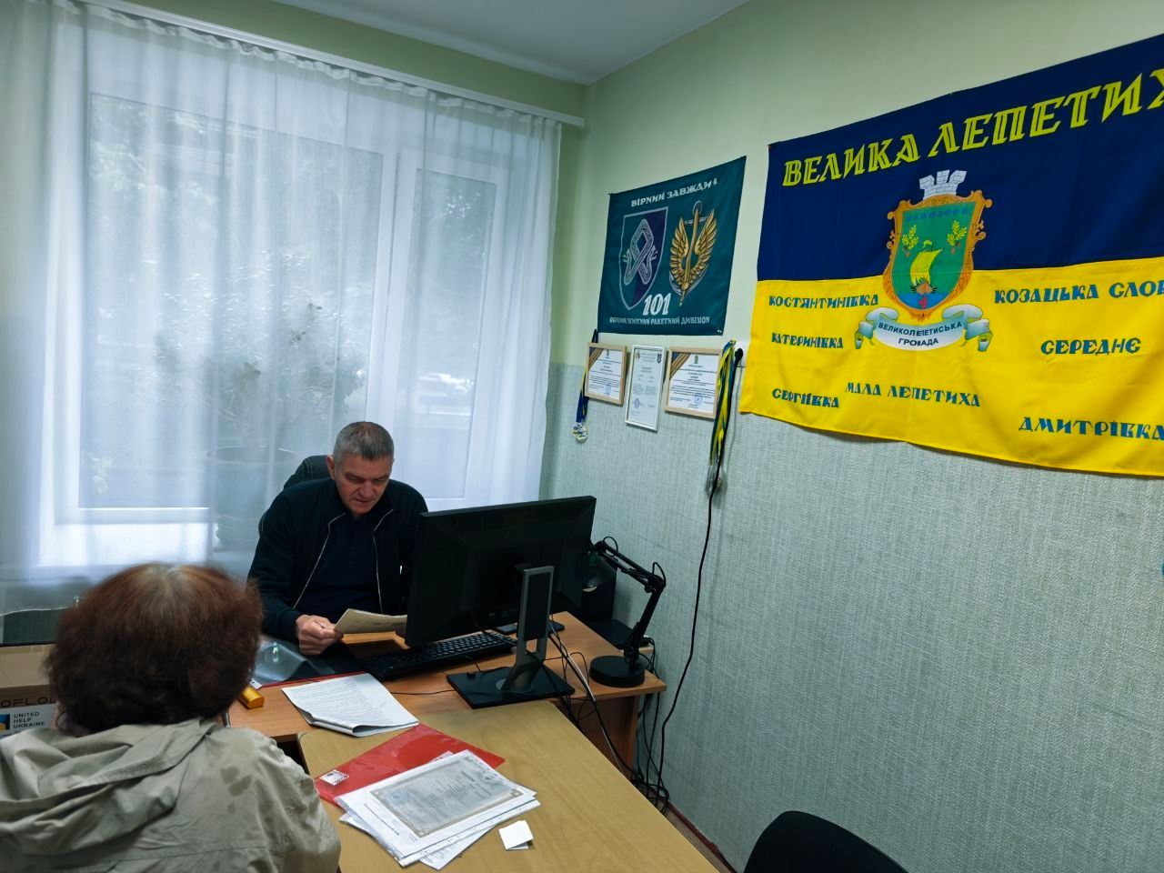 Чоловік-консультант у темній спортивній кофті сидить за робочим столом у кабінеті Координаційного центру «Вільні разом» у Хмельницькому й веде прийом жінки-відвідувачки, надаючи їй необхідні адміністративні послуги та консультації фахівцями відділу ЦНАП. Спеціаліст вивчає паперові документи перед комп'ютерним монітором біля вікна, а на стіні кабінету закріплено темно-синій прапор 101-ї окремої бригади охорони ГШ ЗСУ та великий жовто-блакитний прапор Великолепетиської громади з офіційним гербом і назвами населених пунктів (Велика Лепетиха, Костянтинівка, Мала Лепетиха, Дмитрівка, Сергіївка), що ілюструє процес успішного опрацювання 34 звернень для забезпечення правових і соціальних потреб переселенців із Херсонщини в евакуації.