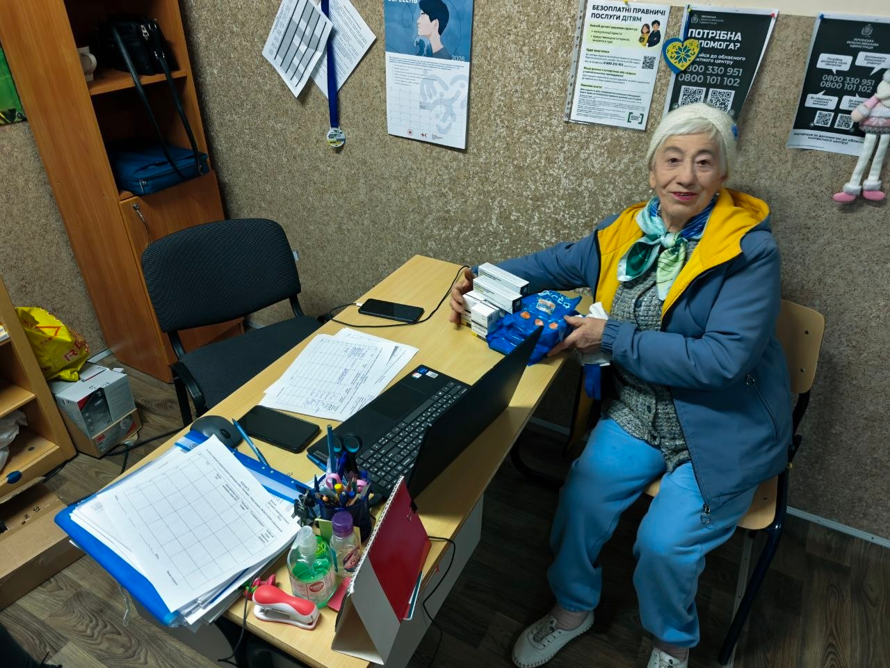 Літня жінка-відвідувачка у блакитному костюмі та жовто-синій куртці сидить за робочим столом у кабінеті Координаційного центру «Вільні разом» у Хмельницькому, отримуючи гуманітарну допомогу у вигляді лікарських засобів. Перед громадянкою на дерев'яному столі розкладено відкритий ноутбук, офіційні бланки, робочі папери, канцелярію та пляшку з антисептиком, а в руках вона тримає видані медичні препарати. Кадр ілюструє системну підтримку переселенців із Херсонщини, під час якої відвідувачам хабу за день було успішно видано 23 одиниці лікарських засобів, 30 захисних масок та 11 шприців для забезпечення їхніх першочергових медичних потреб в евакуації.