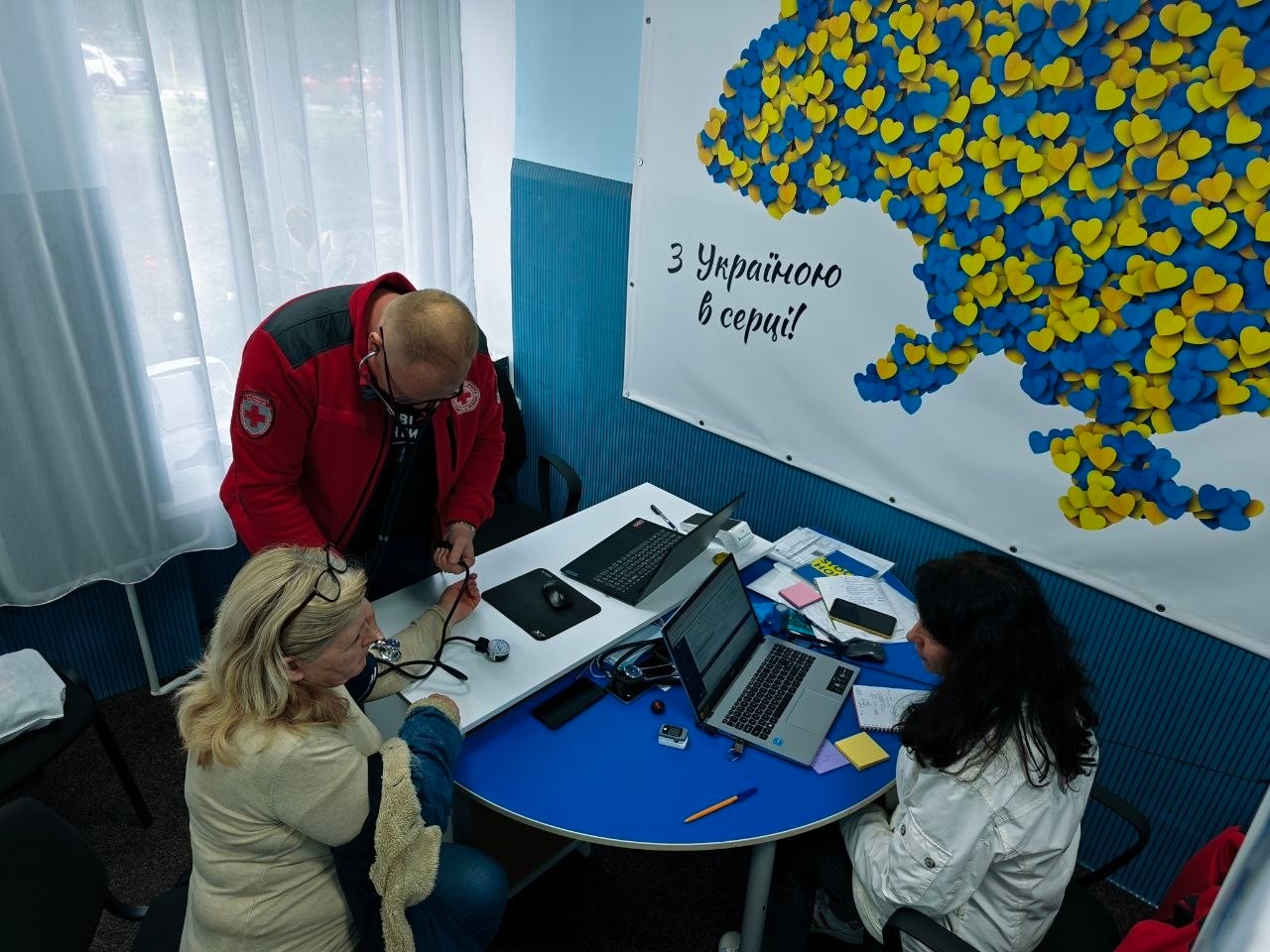 Медичний працівник Товариства Червоного Хреста України у фірмовій червоній куртці вимірює артеріальний тиск літній жінці за робочим столом у кабінеті Координаційного центру «Вільні разом» у Хмельницькому. Поруч за напівкруглим синім столом сидить інша співробітниця хабу перед відкритим ноутбуком, оформлюючи бланки та медичні картки відвідувачів. Прийом проходить на тлі великого білого настінного банера з патріотичною картою України, яка викладена з маленьких синьо-жовтих сердець, та написом «З Україною в серці!», що ілюструє роботу мобільної бригади з надання медичних консультацій та видачі лікарських засобів для 19 громадян із-поміж переселенців із Херсонщини.