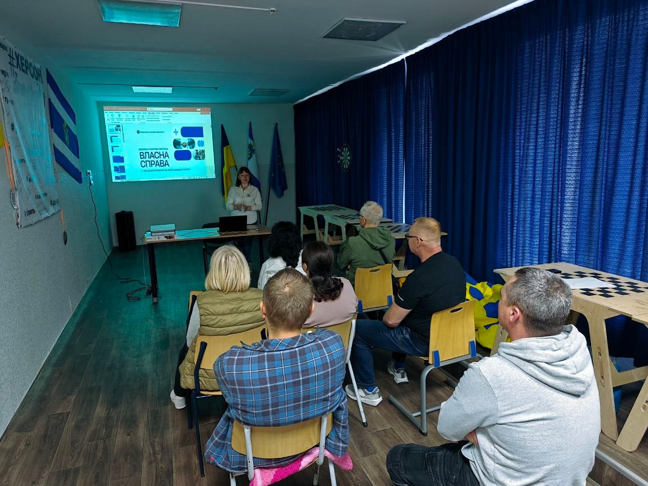 Група переселенців із Херсонщини сидить у залі Координаційного центру «Вільні разом» у Хмельницькому під час зустрічі «Власна справа 2.0: про гранти для старту та масштабування бізнесу». Присутні слухають жінку-лекторку, яка виступає перед ними біля столу з ноутбуком на тлі великого проекційного екрана з презентацією державної грантової програми «Власна справа». Зустріч проходить у просторій залі хабу з темно-синіми шторами, де в кутку встановлено Державний Прапор України, прапор Херсонської області та прапор Європейського Союзу, що ілюструє системну підтримку ВПО у сфері розвитку підприємництва, здобуття фінансової грамотності та економічної адаптації в евакуації.