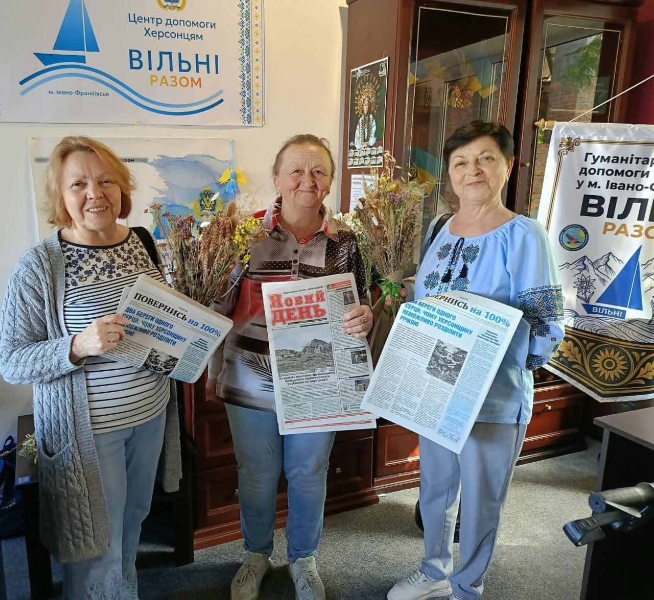 Три жінки похилого віку з-поміж переселенок із Херсонщини радісно позують у приміщенні хабу в Івано-Франківську, тримаючи в руках свіжі примірники друкованої преси. Жінка ліворуч у сірому кардигані та жінка праворуч у блакитній вишиванці демонструють спецвипуск «Повернись на 100%» із заголовком «Два береги одного серця: чому Херсонщину неможливо розділити річкою», а жінка по центру тримає обласну газету «Новий день». Учасниці заходу зафіксовані на тлі офіційного банера з написом «Центр допомоги Херсонцям ВІЛЬНІ РАЗОМ м. Івано-Франківськ», що ілюструє комплексну підтримку земляків в евакуації, у межах якої 18 переселенців отримали примірники рідних видань для збереження єдиного інформаційного простору.