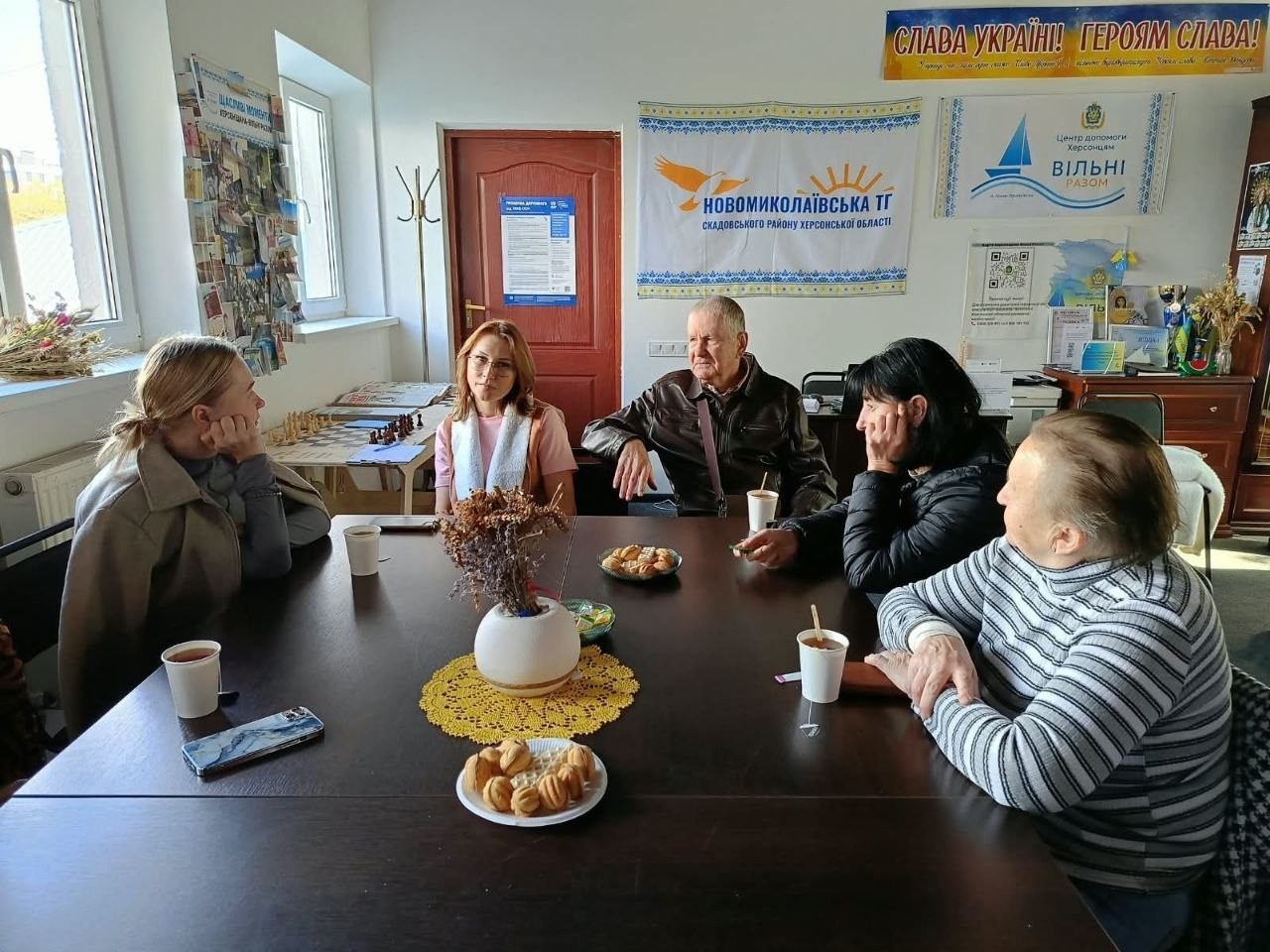 Четверо відвідувачів та спеціалістка затишно спілкуються навколо великого темного столу під час групового психологічного заняття «Моя історія — моя сила» в Координаційному центрі «Вільні разом» в Івано-Франківську. Учасники зустрічі з-поміж переселенців із Херсонщини сидять за чаюванням із солодощами у світлій кімнаті з великими вікнами, де на столі стоять паперові стаканчики, тарілки з печивом і ваза з сухоцвітами на круглій жовтій серветці. Зустріч проходить на тлі великих настінних банерів із написами «Новомиколаївська ТГ Скадовського району Херсонської області» та «Центр допомоги Херсонцям ВІЛЬНІ РАЗОМ м. Івано-Франківськ», що ілюструє реалізацію проєкту «Код Херсонщини: пам’ять, культура, єдність», до якого загалом долучилися 15 учасників для психоемоційної підтримки та згуртування земляків в евакуації.