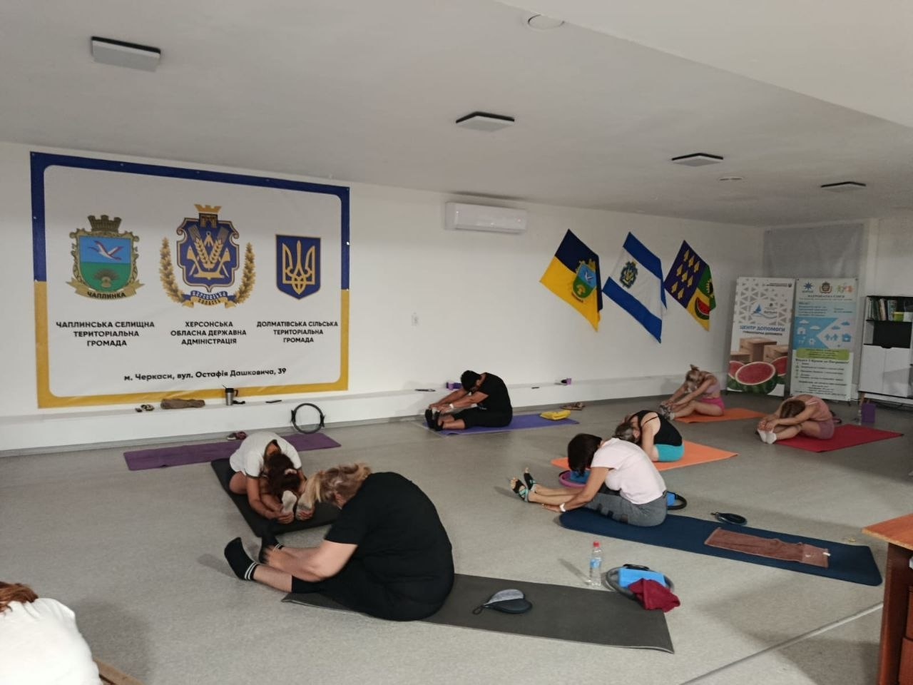 Група жінок різного віку з-поміж переселенок із Херсонщини зосереджено займається спортивною гімнастикою та розтяжкою, сидячи на індивідуальних килимках у просторому залі Координаційного центру «Вільні разом» у Черкасах. Сім учасниць тренування у спортивному одязі синхронно виконують нахили тулуба вперед до ніг у світлій кімнаті з кондиціонером. Ліворуч на стіні встановлено великий інформаційний банер із гербами Чаплинської селищної, Долматівської сільської громад та Херсонської ОДА, а праворуч у кутку закріплено три патріотичні прапори, що ілюструє проведення занять із зумби та йоги для підтримки фізичного й емоційного стану громадян в евакуації.