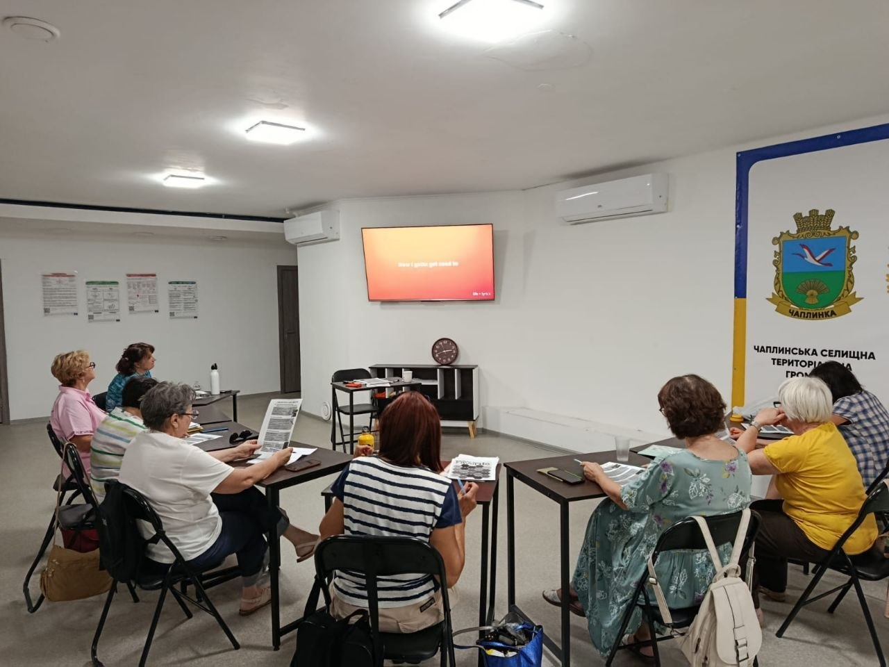 Група жінок старшого віку з-поміж переселенок із Херсонщини та спікерка сидять за партами у просторій залі Координаційного центру «Вільні разом» у Черкасах під час інтерактивного заняття з англійської мови, присвяченого темі музики. Вісім учасниць зустрічі розмістилися навколо столів перед великим настінним телевізійним екраном, де на помаранчево-червоному тлі слайда презентації виведено англійську навчальну фразу «how i am gonna need to». Учеgeneric зафіксовані під час засвоєння тематичної лексики, назв музичних інструментів та закріплення граматики на практиці за допомогою інформаційних друкованих буклетів на тлі великого офіційного банера з гербом та написом «ЧАПЛИНСЬКА СЕЛИЩНА ТЕРИТОРІАЛЬНА ГРОМАДА», що ілюструє комплексну освітню й соціальну підтримку громадян в евакуації.