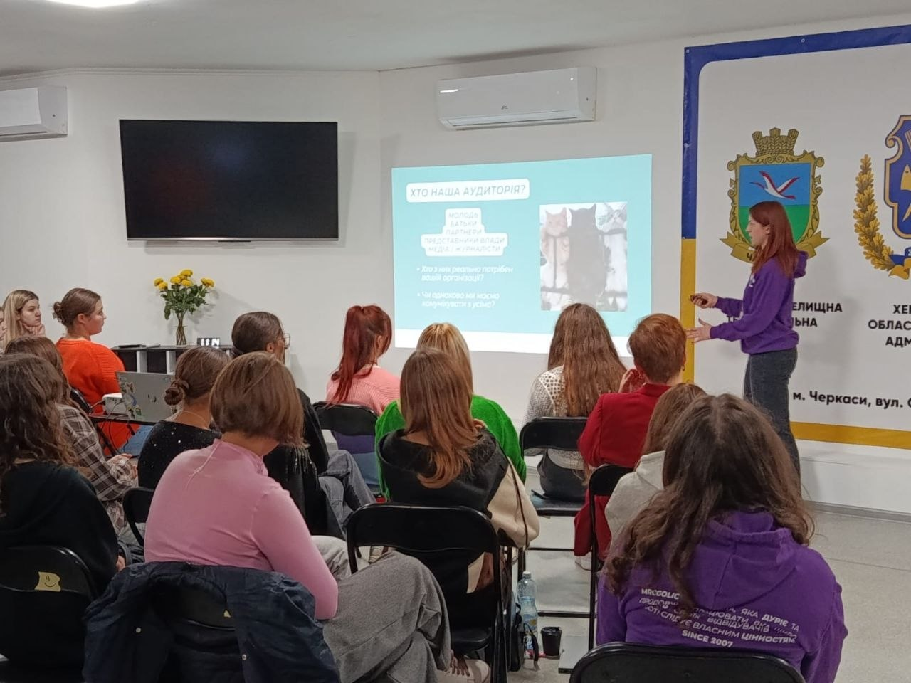 Спікерка у фіолетовій кофті виступає перед великою аудиторією переселенців із Херсонщини в Черкаському координаційному центрі «Вільні разом» під час арттерапевтичної майстерки «Моя кольорова осінь» із практиками емоційного розвантаження. Близько п'ятнадцяти дівчат та жінок різного віку сидять на стільцях у просторій залі, зосереджено слухаючи лекцію та роздивляючись слайд презентації на проекційному екрані із заголовком «ХТО НАША АУДИТОРІЯ?». Зустріч проходить у світлому приміщенні з кондиціонерами на тлі великого офіційного банера з гербом та підписом «ХЕРСОНСЬКА ОБЛАСНА ВІЙСЬКОВА АДМІНІСТРАЦІЯ м. Черкаси», що ілюструє системну діяльність хабу з надання психоемоційної підтримки родинам в евакуації, організації їхнього корисного дозвілля та всебічної соціальної адаптації.