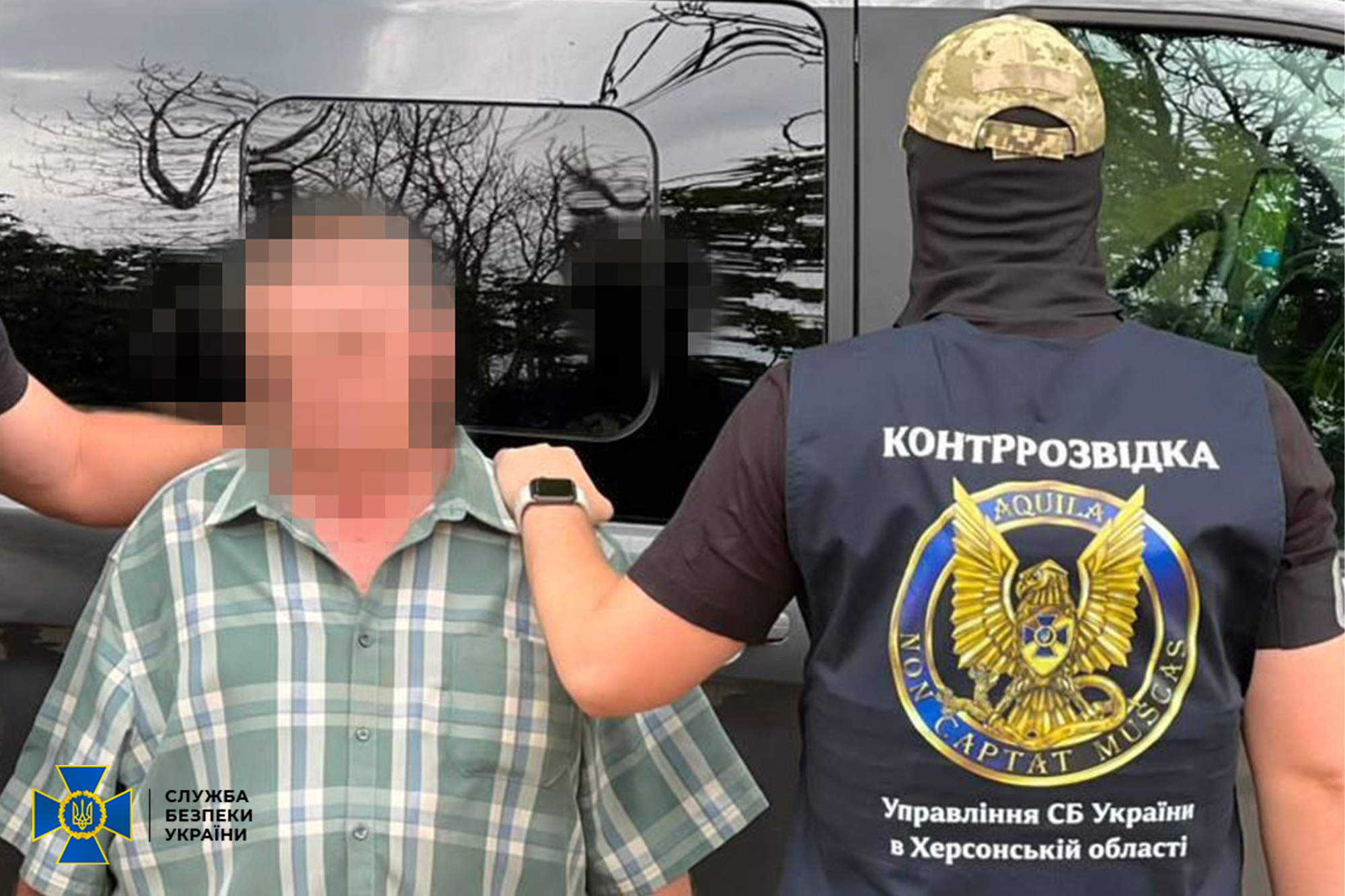 Співробітник контррозвідки Служби безпеки України у тактичній камуфляжній кепці та балаклаві затримує ворожого агента біля автомобіля, тримаючи його за плече під час офіційної безпекової операції у Херсоні. Спецназівець зафіксований зі спини у фірмовому темно-синьому жилеті з великими білими написами «КОНТРРОЗВІДКА» та «Управління СБ України в Херсонській області», по центру якого розміщено круглу емблему підрозділу із золотим соколом та латинським девізом «Aquila non captat muscas». Ліворуч на передньому плані стоїть затриманий чоловік у зеленій кардигані у клітинку з розмитим (пікселізованим) обличчям, а у лівому нижньому кутку кадру нанесено офіційний логотип Служби безпеки України з тризубом, що ілюструє затримання контррозвідкою колишнього авіатехніка ЗСУ, який під час окупації міста увійшов до складу фсб, тривалий час перебував у «сплячому» режимі, а згодом коригував удари КАБами, дронами та артилерією по оборонних об’єктах обласного центру.