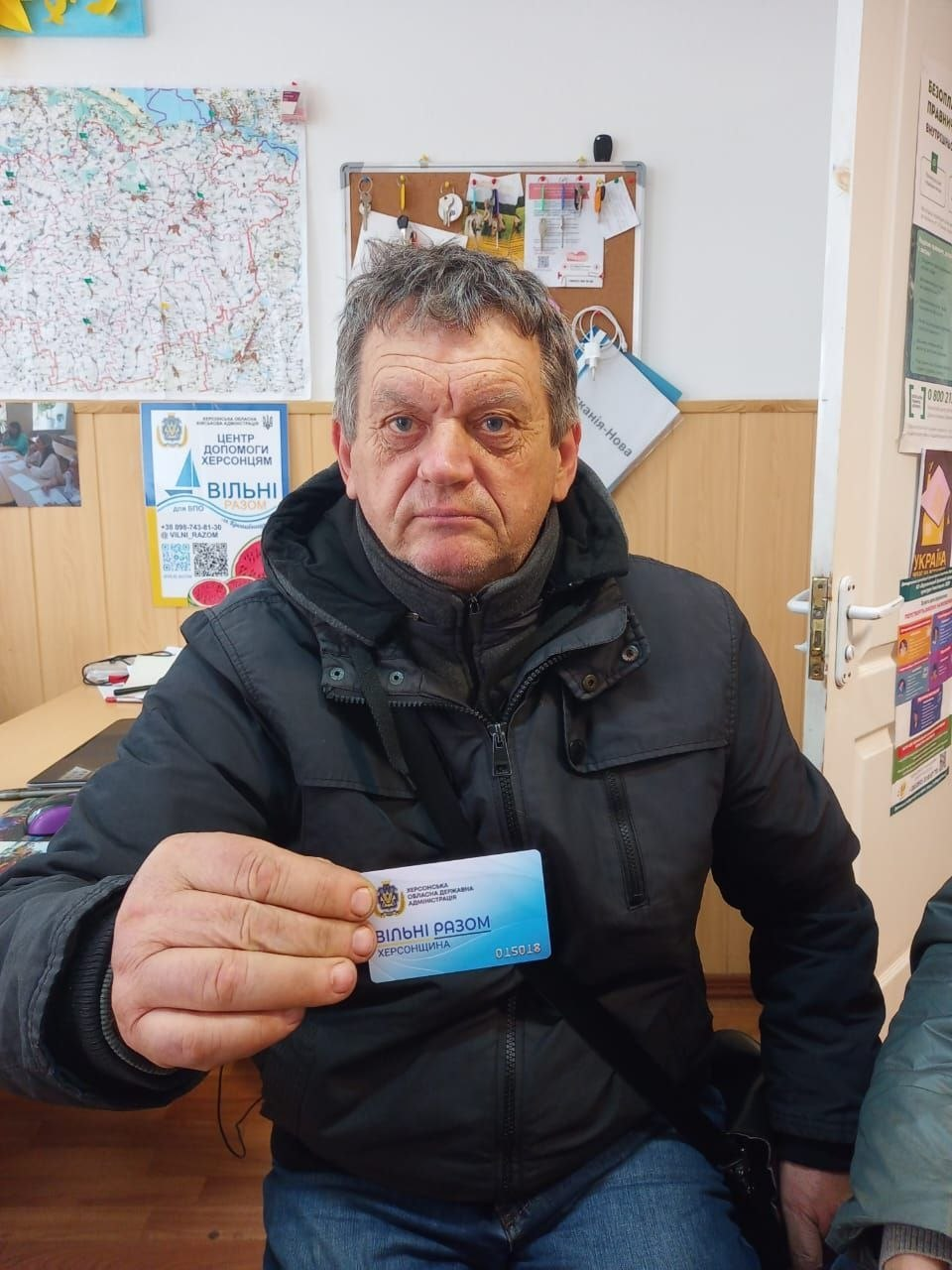 Чоловік середнього віку з-поміж переселенців із Херсонщини сидить у кабінеті Координаційного центру «Вільні разом» у Кропивницькому та демонструє перед собою отриману пластикову «Картку Херсонщини» з бірюзово-блакитним патріотичним дизайном. Відвідувач у теплій чорній зимовій куртці тримає картку в правій руці, де чітко видно офіційний логотип Херсонської обласної державної адміністрації, великий напис «ВІЛЬНІ РАЗОМ ХЕРСОНЩИНА» та унікальний серійний номер 015018. На задньому плані на стіні зафіксовано географічну карту України, коркову дошку з бланками та фірмовий плакат хабу «ЦЕНТР ДОПОМОГИ ХЕРСОНЦЯМ ВІЛЬНІ РАЗОМ для ВПО м. Кропивницький», що ілюструє системну реалізацію важливого соціального проєкту, у межах якого нові картки отримали 8 осіб для забезпечення комплексної підтримки та пільг в евакуації.