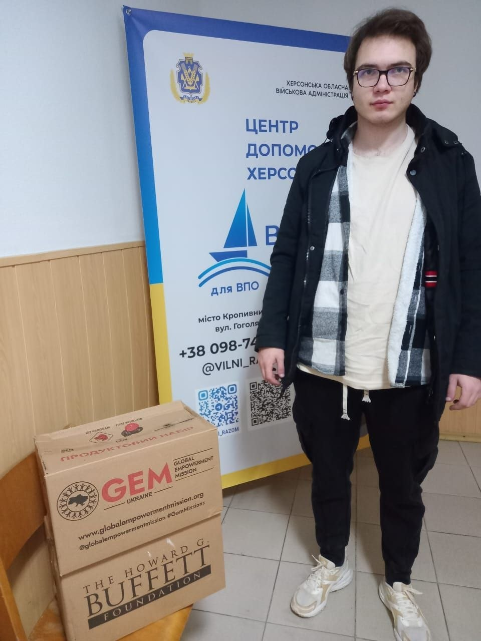 Молодий чоловік в окулярах, чорній куртці та кардигані у клітинку стоїть у приміщенні Координаційного центру «Вільні разом» у Кропивницькому поруч із великою картонною коробкою гуманітарної допомоги. Хлопець із-поміж новоприбулих переселенців із Херсонщини зафіксований у повний зріст на тлі великого офіційного вертикального банера з гербом ХОДА та написом «ЦЕНТР ДОПОМОГИ ХЕРСОНЦЯМ ВІЛЬНІ РАЗОМ для ВПО місто Кропивницький». Ліворуч на підлозі стоїть великий гуманітарний бокс із маркуванням міжнародних благодійних організацій «GEM (Global Empowerment Mission)» та «The Howard G. Buffett Foundation», на верхній частині якого нанесено великий червоний напис «ПРОДУКТОВИЙ НАБІР», що ілюструє процес забезпечення 5 родин новоприбулих ВПО базовими продовольчими пайками для їхньої першочергової підтримки в евакуації.