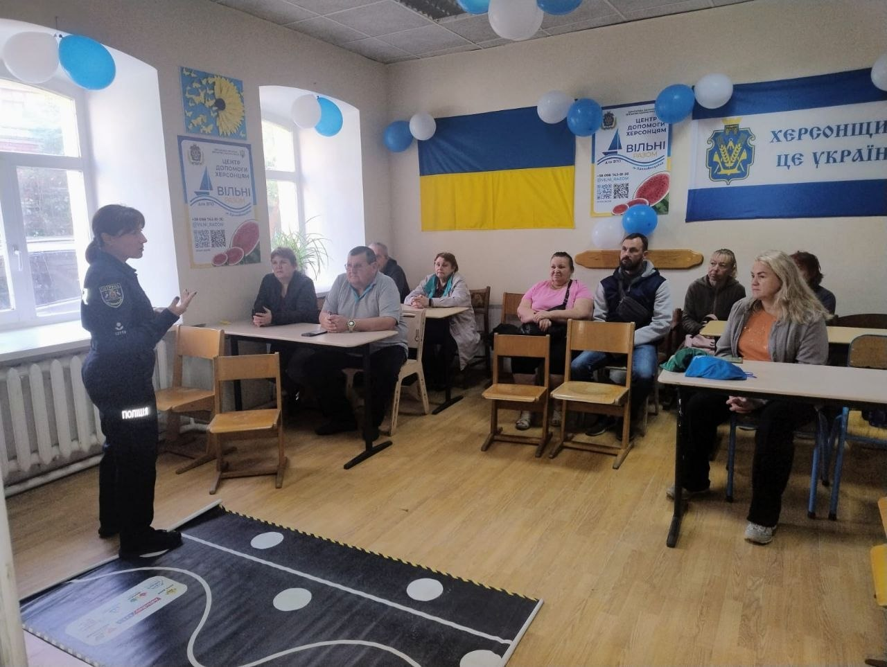 Співробітниця патрульної поліції виступає перед групою переселенців із Херсонщини в залі Координаційного центру «Вільні разом» у Кропивницькому під час практичного інтерактивного заняття з правил дорожнього руху. Близько десяти відвідувачів хабу уважно слухають лекторку, сидячи за навчальними партами у світлій кімнаті біля вікон. На передньому плані на підлозі розстелено великий ігровий чорний килимок-тренажер із білою дорожньою розміткою для моделювання дорожніх ситуацій. Зустріч проходить на тлі великого Державного Прапора України, прапора Херсонської області із написом «Херсонщина — це Україна» та офіційного банера «ЦЕНТР ДОПОМОГИ ХЕРСОНЦЯМ ВІЛЬНІ РАЗОМ», що ілюструє спільну правопросвітницьку та безпекову діяльність хабу й поліції для підтримки 10 учасників з-поміж ВПО в евакуації.
