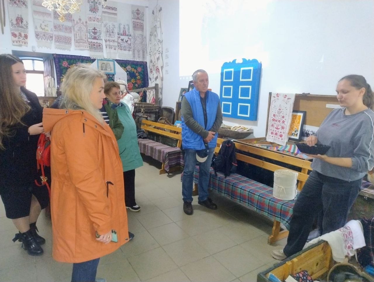 Група переселенців із Херсонщини та екскурсоводка спілкуються в автентичній залі етнолабораторії «Баба Єлька» у Кропивницькому під час екскурсії з майстер-класом із виконання традиційних українських весільних пісень. Праворуч біля дерев'яної лави стоїть молода майстриня-лекторка у сірій кофті, яка тримає в руках старовинний побутовий предмет, а навпроти неї розмістилися відвідувачі хабу, серед яких виділяється жінка в помаранчевій куртці на передньому плані та чоловік у синьому жилеті. Зустріч проходить у затишному музейному просторі, де стіни рясно прикрашені колекціями автентичних вишитих рушників, тканими доріжками, сорочками та предметами українського народного побуту, що ілюструє соціокультурну інтеграцію та психоемоційну підтримку херсонців в евакуації, організовану командою Координаційного центру «Вільні разом».