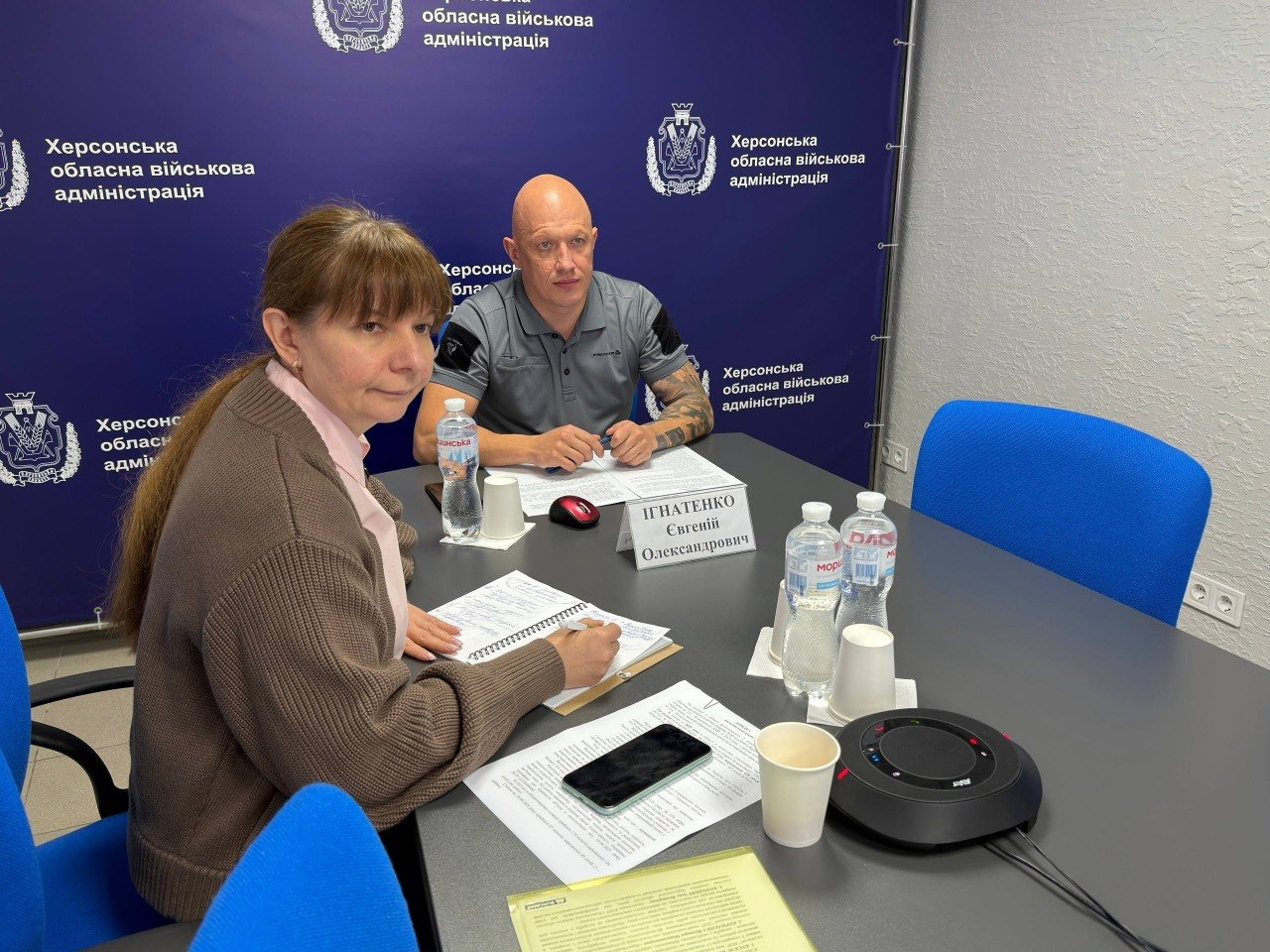 Заступник начальника Херсонської ОВА Євгеній Ігнатенко та директорка Департаменту здоров'я ХОДА Тетяна Карчевич сидять за робочим столом у кабінеті під час онлайн-участі у селекторній нараді під головуванням Міністра охорони здоров’я України Віктора Ляшка. На передньому плані ліворуч Тетяна Карчевич у коричневому трикотажному кардигані робить нотатки ручкою у блокноті, а по центру столу навпроти неї сидить Євгеній Ігнатенко у сірій футболці-поло з піднятими руками. Перед посадовцями на довгому темно-сірому столі розкладено робочі папери, бланки, мобільний телефон, пластикові пляшки з водою, паперові стаканчики та встановлено круглий чорний спікерфон конференц-зв'язку. Зустріч проходить на тлі великого темно-синього офіційного банера з чисельними повтореннями герба ХОВА та білими написами «Херсонська обласна військова адміністрація», що ілюструє обговорення готовності захисних споруд і підземних пологово-операційних блоків у медзакладах Херсонщини для забезпечення безпеки породіль та новонароджених.