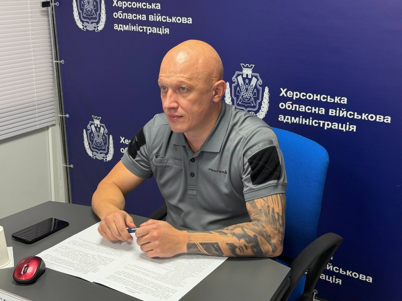 Заступник начальника Херсонської ОВА Євгеній Ігнатенко сидить за робочим столом у кабінеті, беручи участь у селекторній нараді під головуванням Міністра охорони здоров’я України Віктора Ляшка. Чоловік у сірій тактичній футболці-поло з темними велкро-панелями на рукавах показаний крупним планом, коли він вивчає розкладені робочі папери та тримає в руках синю кулькову ручку. На сірому столі ліворуч від нього лежать мобільний телефон та комп'ютерна миша з червоною вставкою. Посадовець розмістився на яскраво-блакитному стільці на тлі великого темно-синього офіційного банера з чисельними повтореннями герба ХОВА та білими написами «Херсонська обласна військова адміністрація», що ілюструє обговорення готовності захисних споруд і підземних пологово-операційних блоків для безпеки матерів і немовлят.