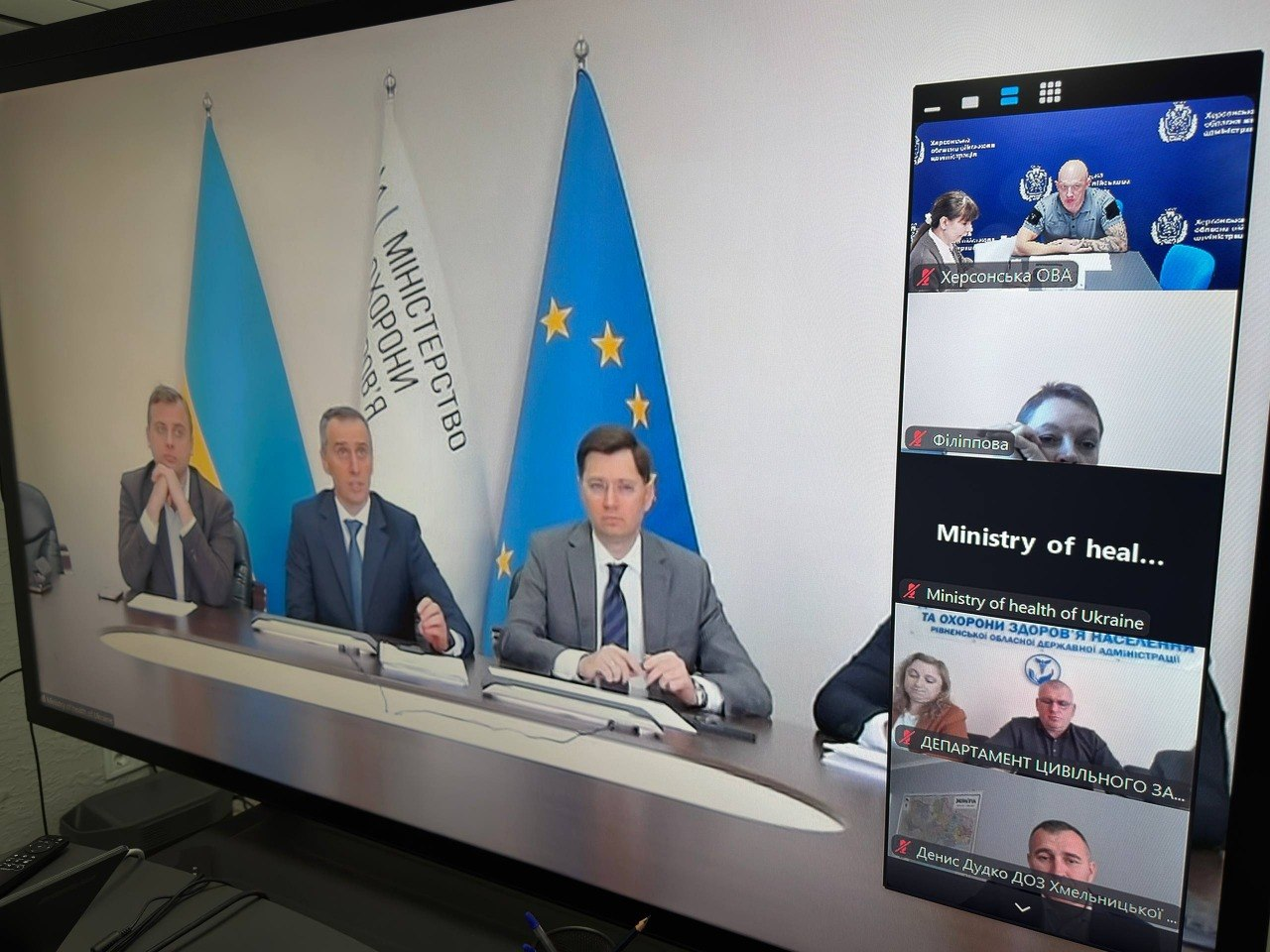 Великий монітор у конференц-залі відображає трансляцію селекторної наради під головуванням Міністра охорони здоров’я України Віктора Ляшка, присвяченої безпеці породіль та новонароджених. На основній частині екрана видно засідання керівництва Міністерства охорони здоров'я України на тлі Державного Прапора України, прапора Європейського Союзу та білого прапора МОЗ. Праворуч у вертикальному стовпчику вікон онлайн-зв'язку транслюються інші учасники наради, серед яких у верхньому віконці з підписом «Херсонська ОВА» перебувають заступник начальника Херсонської ОВА Євгеній Ігнатенко та директорка Департаменту здоров'я ХОДА Тетяна Карчевич. Нижче розташовані вікна підключень інших відомств та регіонів, що ілюструє міжрегіональне обговорення готовності захисних споруд та облаштування підземних пологово-операційних блоків у лікарнях.