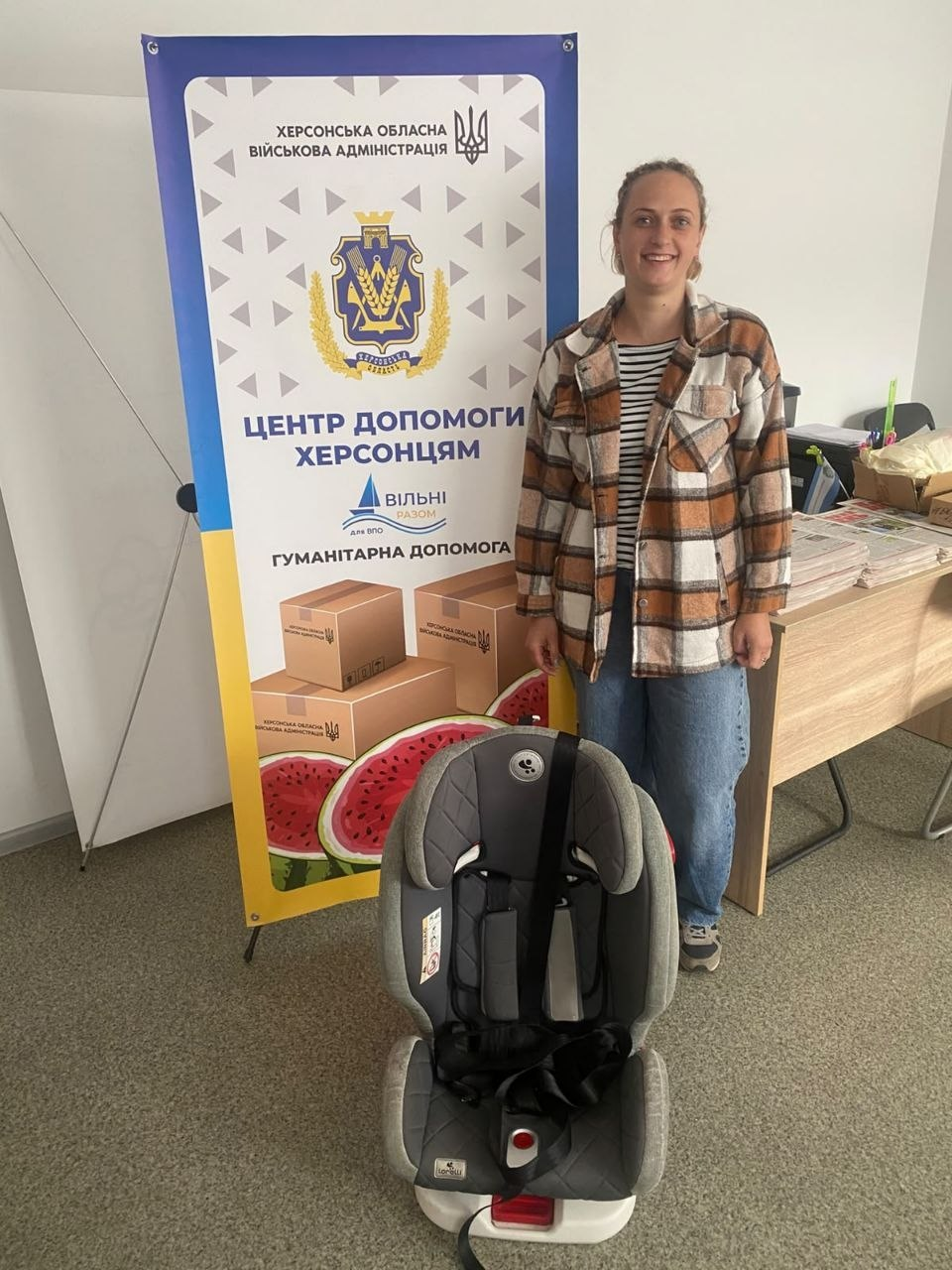 Молода жінка у картатій сорочці та джинсах радісно посміхається у приміщенні Координаційного центру «Вільні разом» у Житомирі, отримавши гуманітарну допомогу у вигляді дитячого автокрісла від волонтерів. Відвідувачка з-поміж переселенок із Херсонщини стоїть біля робочого столу на тлі великого фірмового банера з офіційним гербом ХОВА та написами «ХЕРСОНСЬКА ОБЛАСНА ВІЙСЬКОВА АДМІНІСТРАЦІЯ», «ЦЕНТР ДОПОМОГИ ХЕРСОНЦЯМ ВІЛЬНІ РАЗОМ для ВПО» та «ГУМАНІТАРНА ДОПОМОГА», на якому також зображені коробки та скибки херсонського кавуна. Попереду неї на підлозі встановлено надійне дитяче автомобільне крісло сірого кольору з чорними пасками безпеки, що ілюструє системну підтримку евакуйованих родин із дітьми для забезпечення їхнього комфорту та безпеки під час поїздок.