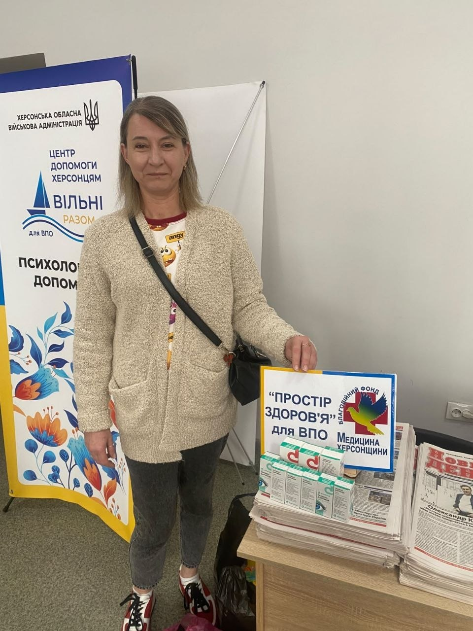 Жінка середнього віку у світлій в'язаній кофті та джинсах стоїть у приміщенні Координаційного центру «Вільні разом» у Житомирі поруч із робочим столом, де розкладено гуманітарну допомогу у вигляді лікарських засобів та друкованої преси від БО БФ «Медицина Херсонщини». Відвідувачка з-поміж переселенок із Херсонщини зафіксована у повний зріст на тлі великого фірмового банера хабу з офіційним гербом ХОВА та написами «ЦЕНТР ДОПОМОГИ ХЕРСОНЦЯМ ВІЛЬНІ РАЗОМ для ВПО» та «ПСИХОЛОГІЧНА ДОПОМОГА». Вона тримає руку на табличці з написом «"ПРОСТІР ЗДОРОВ'Я" для ВПО Медицина ХЕРСОНЩИНИ БЛАГОДІЙНИЙ ФОНД», яка встановлена на столі поверх стосів обласної газети «Новий день» поруч із медичними препаратами, що ілюструє системну турботу про здоров'я та інформаційну підтримку громадян в евакуації.