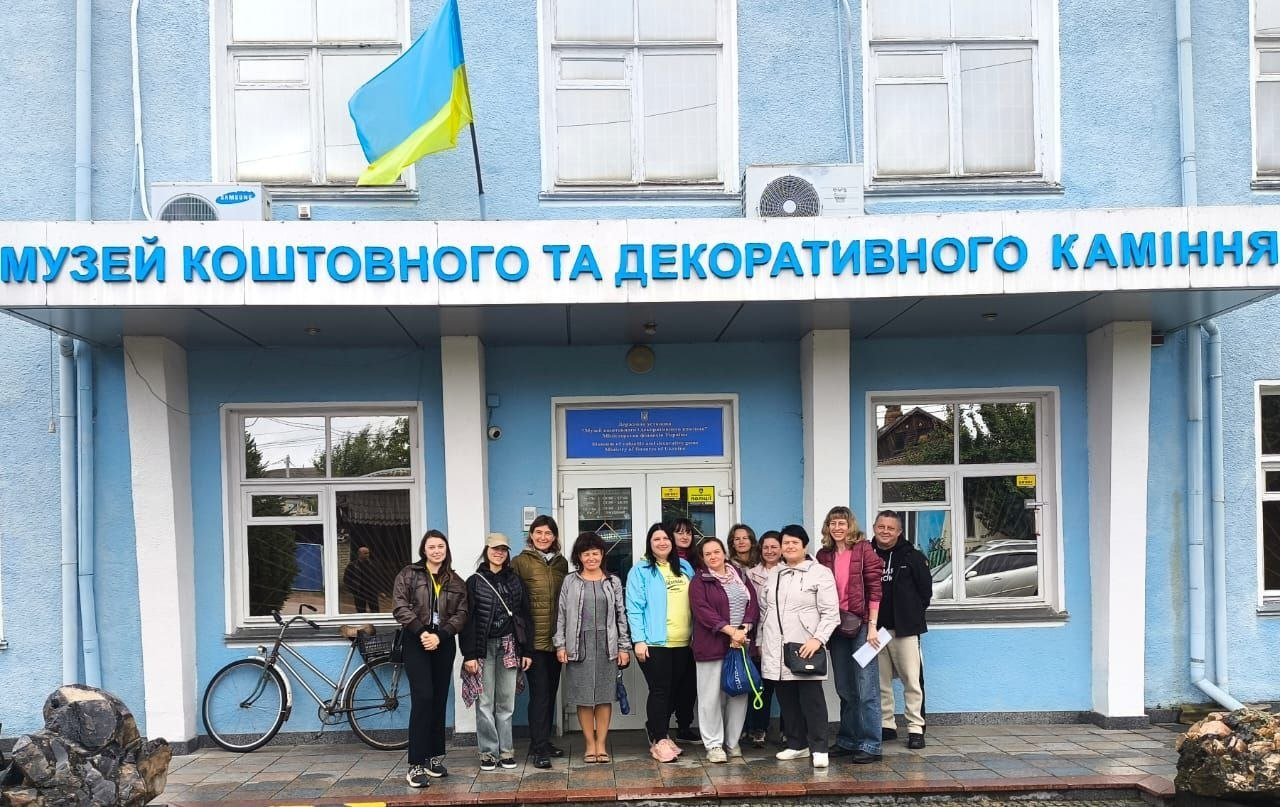 Група переселенців із Херсонщини разом із супроводжуючими вишикувалися для спільного фото біля центрального входу до Музею коштовного та декоративного каміння у смт Хорошів під час виїзної екскурсії. Екскурсанти у повсякденному осінньому одязі та куртках стоять на брукованому майданчику під великою синьо-білою вивіскою закладу на тлі блакитного фасаду будівлі, де над під'їздом встановлено Державний Прапор України. Ліворуч біля вікна припарковано міський велосипед, а на передньому плані видно великі декоративні валуни гірських порід, що ілюструє соціокультурну поїздку для херсонців у Житомирській області, організовану Координаційним центром «Вільні разом» за сприяння БФ «Рокада», який забезпечив групу комфортним транспортом задля їхньої психоемоційної підтримки та соціальної адаптації в евакуації.