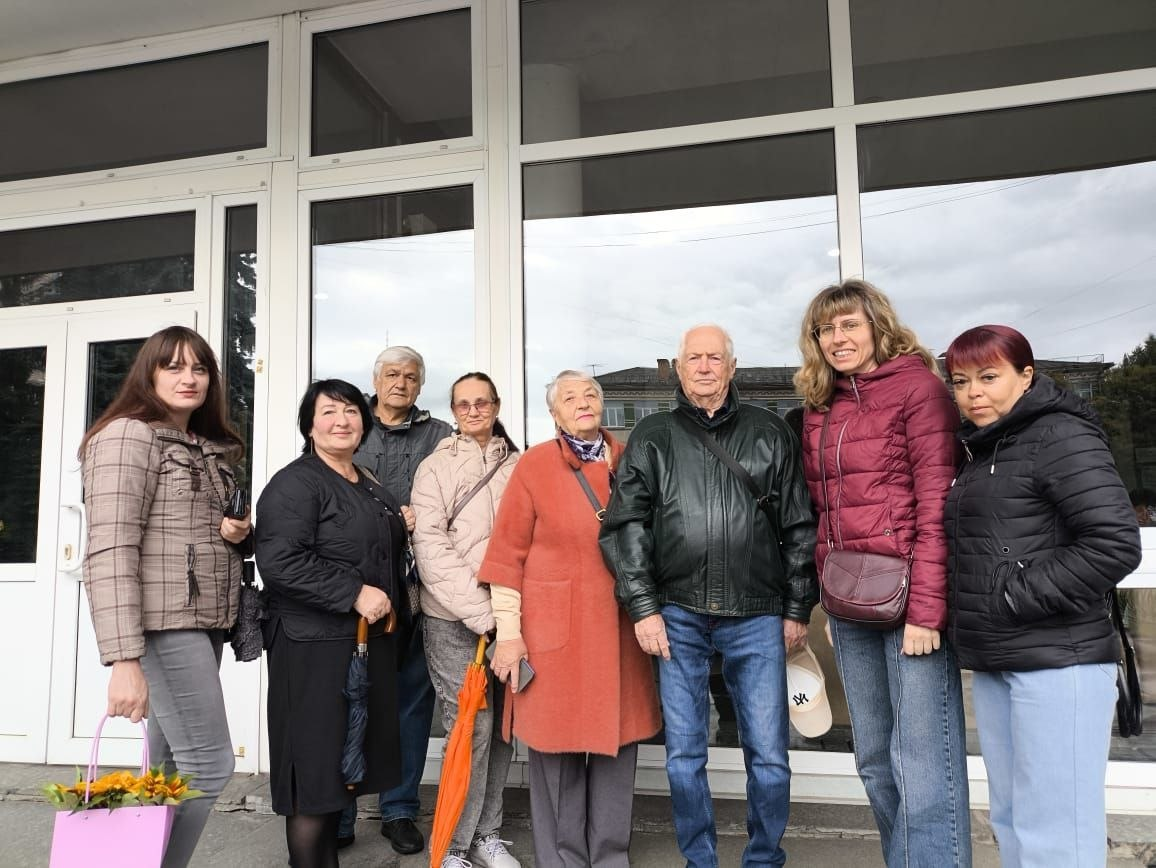 Група переселенців із Херсонщини разом із супроводжуючими дружньо позує для спільного фото біля скляного входу до будівлі житомирського телецентру перед початком або після завершення культурного заходу. Відвідувачі Координаційного центру в осінньому повсякденному одязі та куртках посміхаються на тлі великих панорамних вікон, у відображенні яких видно міські будинки та хмарне небо, а жінка крайня ліворуч тримає в руках подарунковий пакунок із квітами. Кадр ілюструє організацію культурного дозвілля для громадян в евакуації, які відвідали концерт гурту «Лос Янковерс» у межах системної програми психоемоційної підтримки та соціальної адаптації херсонців, що реалізується командою Координаційного центру «Вільні разом» у Житомирі.