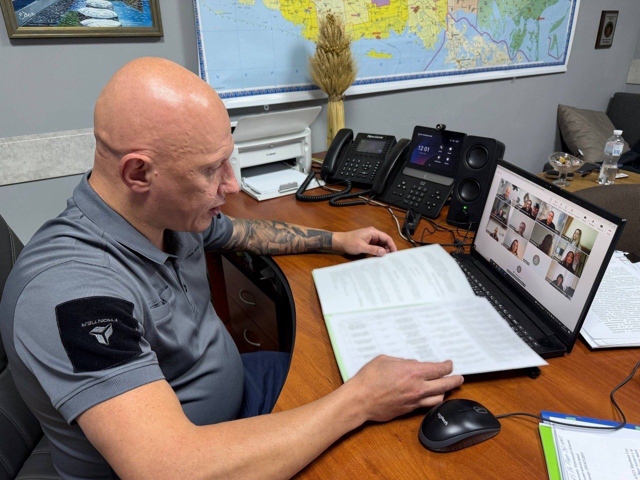 Заступник начальника Херсонської ОВА Євгеній Ігнатенко сидить за своїм робочим столом в офісному кабінеті під час проведення засідання обласної комісії у форматі онлайн-конференції. Чоловік у сірій тактичній футболці-поло показаний крупним планом у профіль, коли він вивчає розгорнуту велику папку з документами та веде діалог із учасниками зустрічі через відкритий ноутбук. На дерев'яному столі перед ним розташовані комп'ютерна миша, стаціонарні телефони, апарат селекторного зв'язку, а на задньому плані видно офісну техніку та велику карту на стіні. Кадр ілюструє ухвалення важливого рішення щодо надання одноразової матеріальної допомоги коштом обласного бюджету, за результатами якого розглянули 23 заяви та призначили виплати сім'ям полеглих воїнів із Херсонщини на загальну суму 1 мільйон 150 тисяч гривень.