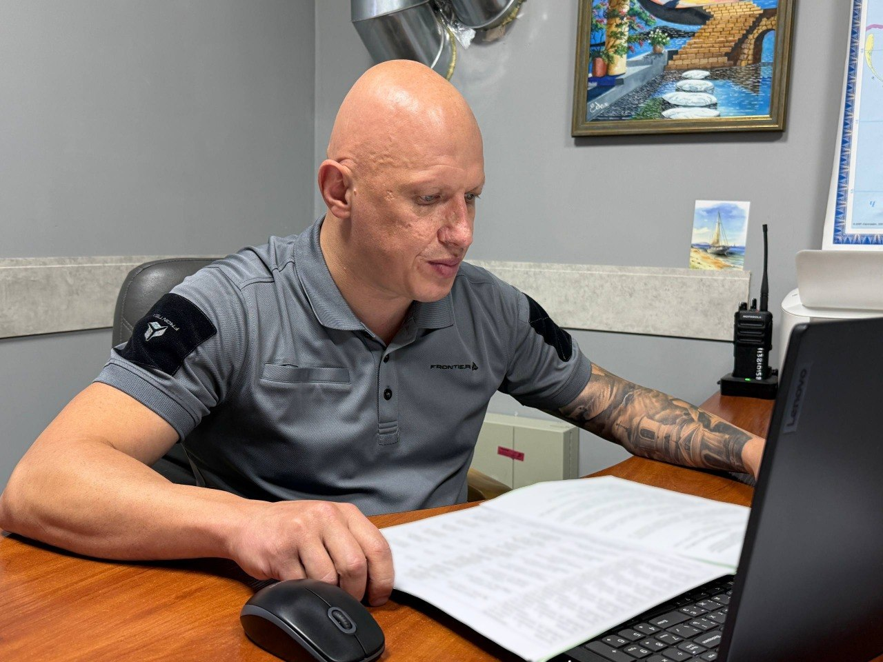 Заступник начальника Херсонської ОВА Євгеній Ігнатенко сидить за своїм робочим столом в офісному кабінеті під час проведення засідання обласної комісії у форматі онлайн-конференції. Посадовець у сірій тактичній футболці-поло показаний крупним планом у напівпрофіль, коли він зосереджено вивчає розгорнуті документи у папці та звіряє дані з інформацією на екрані ноутбука Lenovo. На дерев'яному столі перед ним розташовані комп'ютерна миша та портативна радіостанція (рація) на зарядній док-станції, а на задньому плані видно світло-сіру стіну з невеликою картиною у рамці, що ілюструє ухвалення рішення про призначення одноразових виплат сім'ям загиблих Захисників і Захисниць України на загальну суму 1 мільйон 150 тисяч гривень за результатами розгляду 23 заяв.