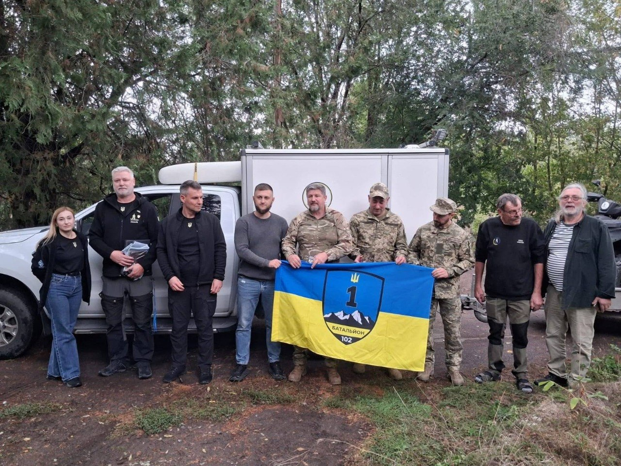 Група чоловіків та жінка разом із військовослужбовцями позують на тлі лісистої місцевості, тримаючи великий жовто-блакитний прапор 1-го батальйону 102-ї окремої бригади Сил територіальної оборони ЗСУ, під час передачі матеріально-технічної допомоги від шведських партнерів для підрозділів на Херсонському напрямку. Дев'ятеро учасників зустрічі у військовому камуфляжі та повсякденному цивільному одязі вишикувалися в ряд на відкритому повітрі біля білого пікапа Ford Ranger та фургона, які громадська організація «Svenskbyborna» разом із обладнанням зв'язку, системами автономного живлення, медичним спорядженням та формою доставила для потреб українських оборонців. На прапорі по центру зображено темно-синій щит із цифрою 1, тризубом та засніженими горами з написом «БАТАЛЬЙОН 102», що ілюструє логістичну підтримку шведських благодійників та згуртованість волонтерів задля успішного виконання бійцями бойових завдань.