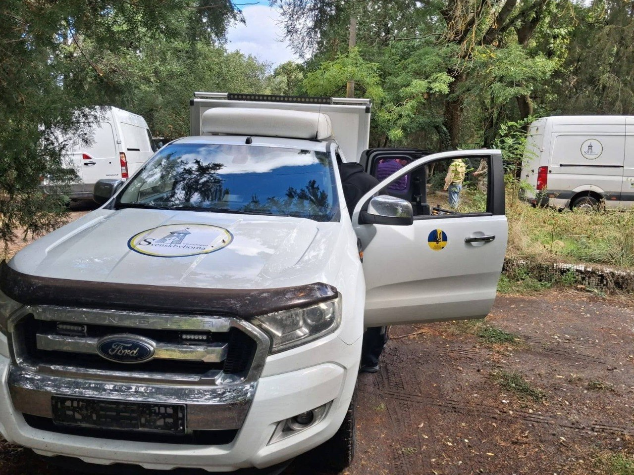 Білий позашляховик Ford Ranger з емблемою шведської громадської організації «Svenskbyborna» на капоті зафіксований крупним планом на лісовій дорозі під час передачі матеріально-технічної допомоги підрозділам Сил оборони на Херсонському напрямку. На капоті та відкритих передніх дверцятах пікапа нанесено фірмові круглі наліпки з малюнком старовинної кірхи та назвою організації, яка передала техніку. Автомобіль стоїть із відкритими пасажирськими дверима, а на задньому плані серед зелених дерев видно інші елементи логістичної партії — білі вантажні фургони Volkswagen Crafter, що ілюструє процес доставки шведськими благодійниками автівок, засобів зв'язку Starlink та автономного живлення для потреб 42-ї окремої бригади морської піхоти та 102-ї окремої бригади ТрО ЗСУ.