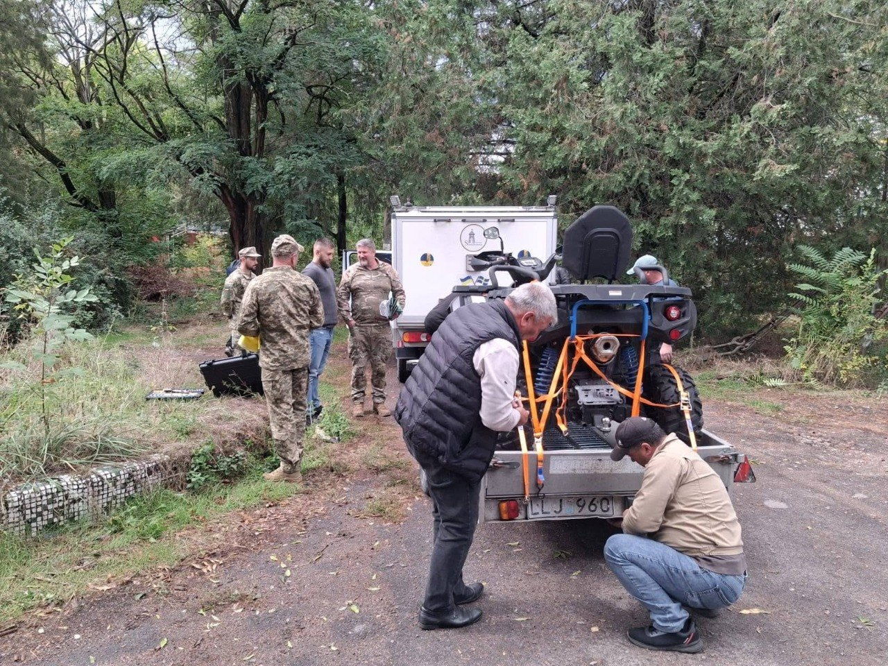 Група чоловіків та військовослужбовців закріплює новий потужний квадроцикл на автомобільному причепі під час передачі матеріально-технічної допомоги від шведських партнерів для підрозділів на Херсонському напрямку. На передньому плані сивочолий чоловік у темному жилеті та його колега навприсядки налаштовують натяжні помаранчеві ремені для фіксації колісної техніки на причепі з іноземним номерним знаком «LLJ 960». На задньому плані біля білого фургона громадської організації «Svenskbyborna» стоять українські військові у камуфляжі ЗСУ та представники волонтерської місії, які обговорюють деталі логістики, що ілюструє процес доставки автомобільної техніки, квадроцикла та обладнання автономного живлення для потреб 42-ї окремої бригади морської піхоти та 102-ї окремої бригади ТрО.