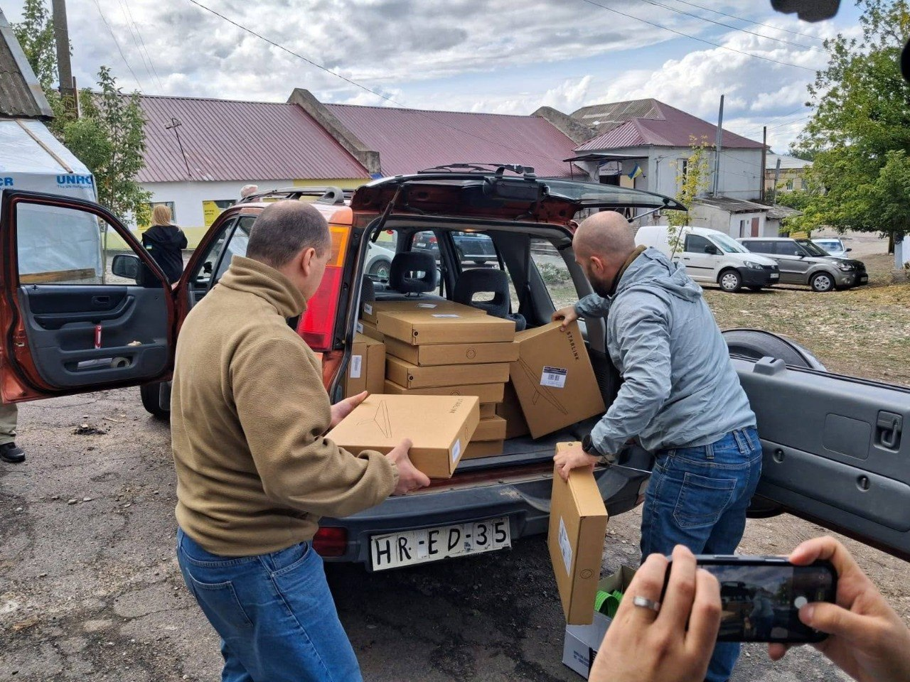 Два чоловіки розвантажують фірмові картонні коробки із терміналами супутникового зв'язку Starlink із багажника автомобіля під час передачі матеріально-технічної допомоги від шведських партнерів для підрозділів на Херсонському напрямку. Чоловік ліворуч у гірчичній флісовій кофті та чоловік праворуч у сірій вітрівці й синіх джинсах акуратно дістають пласкі упакування зі супутниковим обладнанням із заповненого багажного відділення позашляховика. На передньому плані праворуч видно руки іншої людини, яка фіксує процес логістики на камеру смартфона, а на задньому плані відображено сільську вулицю з одноповерховими будівлями, автомобілями та гуманітарним тентом UNHCR, що ілюструє доставку систем зв'язку, автономного живлення та спорядження від громадської організації «Svenskbyborna» для успішного виконання бойових завдань українськими оборонцями.