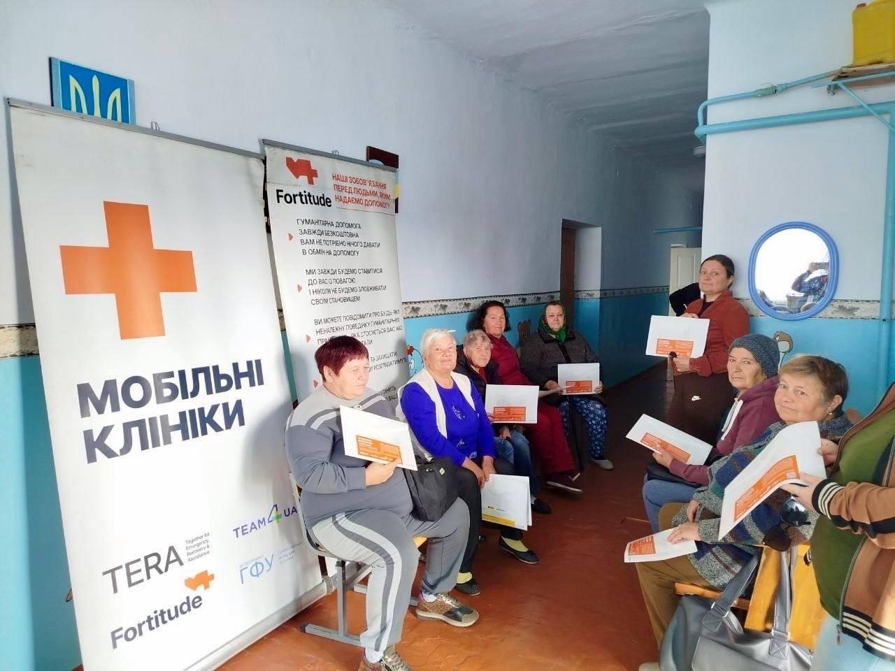 Група жінок та мешканців похилого віку сидить у коридорі приміщення під час виїзного прийому медичної команди «Fortitude» у межах проєкту «Мобільні клініки Півдня», який реалізується на Херсонщині. Ліворуч на передньому плані встановлено великий вертикальний банер із помаранчевим хрестом, великим написом «МОБІЛЬНІ КЛІНІКИ» та логотипами організацій «TERA», «TEAM4UA» і «Fortitude», а поруч розміщено плакат із кодексом поведінки «Наші зобов'язання перед людьми, яким надаємо допомогу». Відвідувачі сидять уздовж стін коридору та уважно вивчають помаранчеві інформаційні буклети в очікуванні консультацій, що ілюструє масштабний виїзд фахівців до десяти населених пунктів області, де понад 410 мешканців безкоштовно отримали обстеження, окуляри та необхідні медикаменти за підтримки Гуманітарного фонду для України (UHF).