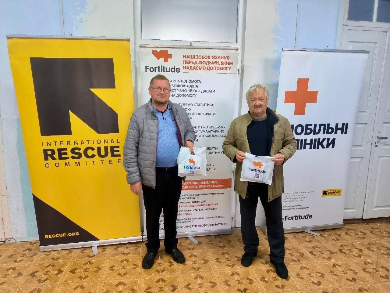 Два чоловіки старшого віку дружньо посміхаються у приміщенні хабу, тримаючи в руках білі тканинні екоторби з помаранчевим логотипом благодійного фонду «Fortitude» під час виїзного прийому медичної команди. Мешканці Херсонщини зафіксовані у повний зріст на тлі офіційних банерів організацій-партнерів: ліворуч встановлено великий яскраво-жовтий банер «INTERNATIONAL RESCUE COMMITTEE», по центру — плакат із кодексом поведінки благодійників «Наші зобов'язання перед людьми, яким надаємо допомогу», а праворуч — вертикальний банер із помаранчевим хрестом та написом «МОБІЛЬНІ КЛІНІКИ». Кадр ілюструє успішне завершення виїздів до десяти населених пунктів області, де понад 410 мешканців безкоштовно отримали консультації лікарів, обстеження, медикаменти та окуляри у межах проєкту «Мобільні клініки Півдня», який реалізується за підтримки Гуманітарного фонду для України (UHF).