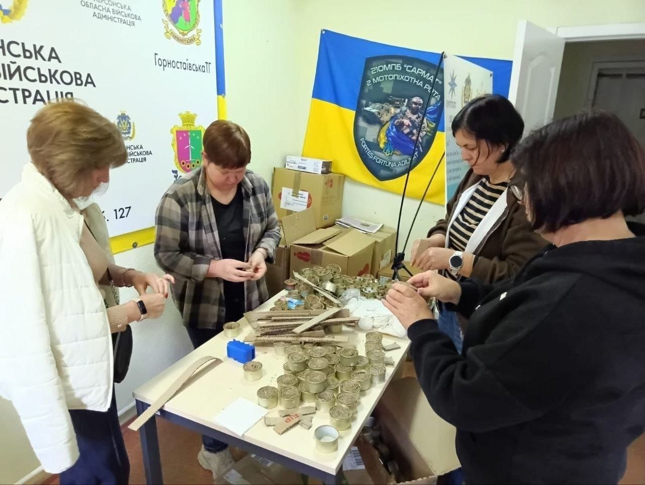 Група жінок з-поміж переселенок із Херсонщини та команди хабу зосереджено працює за великим світлим столом у приміщенні Координаційного центру «Вільні разом» у Дніпрі, виготовляючи окопні свічки та розпалювачі для українських воїнів. Волонтерки скручують смужки гофрованого картону та вкладають їх у порожні бляшанки, яких велика кількість розкладена по центру столу поруч із картонними заготовками та інструментами, готуючи чергову партію до відправлення на фронт. Зустріч проходить у світлій кімнаті на тлі офіційних банерів із гербами Горностаївської громади, Херсонської ОВА та великого синьо-жовтого прапора 21-го окремого мотопіхотного батальйону «Сармат» із гаслом «Fortes fortuna adiuvat», що ілюструє регулярну практичну допомогу евакуйованих громадян Збройним Силам України задля підтримки захисників на передовій.
