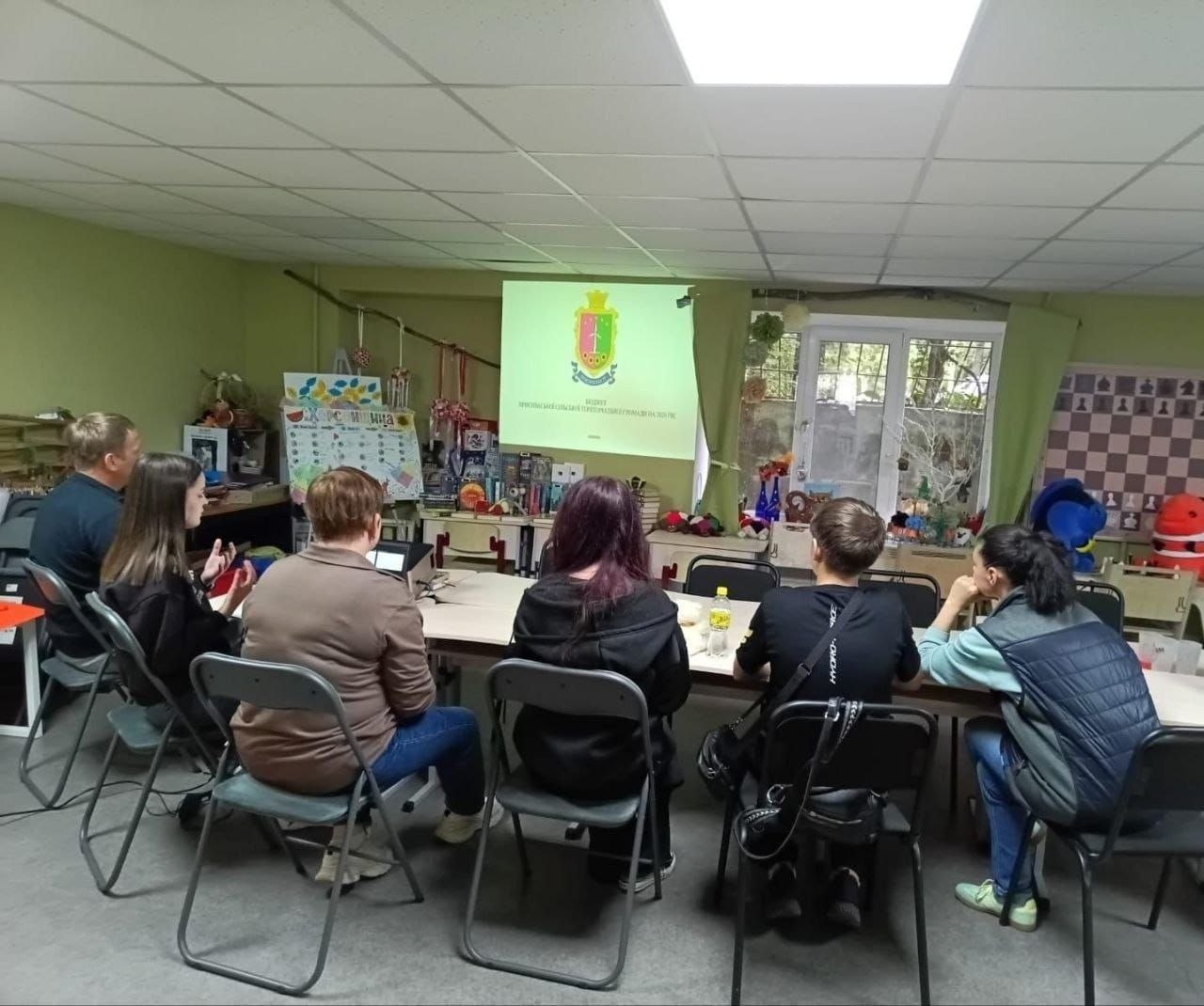 Група переселенців із Херсонщини сидить на стільцях навколо великого столу в затишній кімнаті Координаційного центру «Вільні разом» у Дніпрі під час інформаційно-просвітницької зустрічі щодо принципів формування бюджету громади. Слухачі показані зі спини, коли вони зосереджено спостерігають за проекційним екраном у кінці зали, на якому демонструється офіційний герб Присиваської сільської територіальної громади та стартовий слайд презентації. Захід, який провела представниця Молодіжного конгресу Херсонщини від Присиваської громади, проходить у світлому приміщенні з салатовими стінами, де на задньому плані біля вікна розставлено творчі вироби, книги та дитячий плакат «Херсонщина», що ілюструє залучення громадян в евакуації до правопросвітницьких та інтеграційних програм хабу.