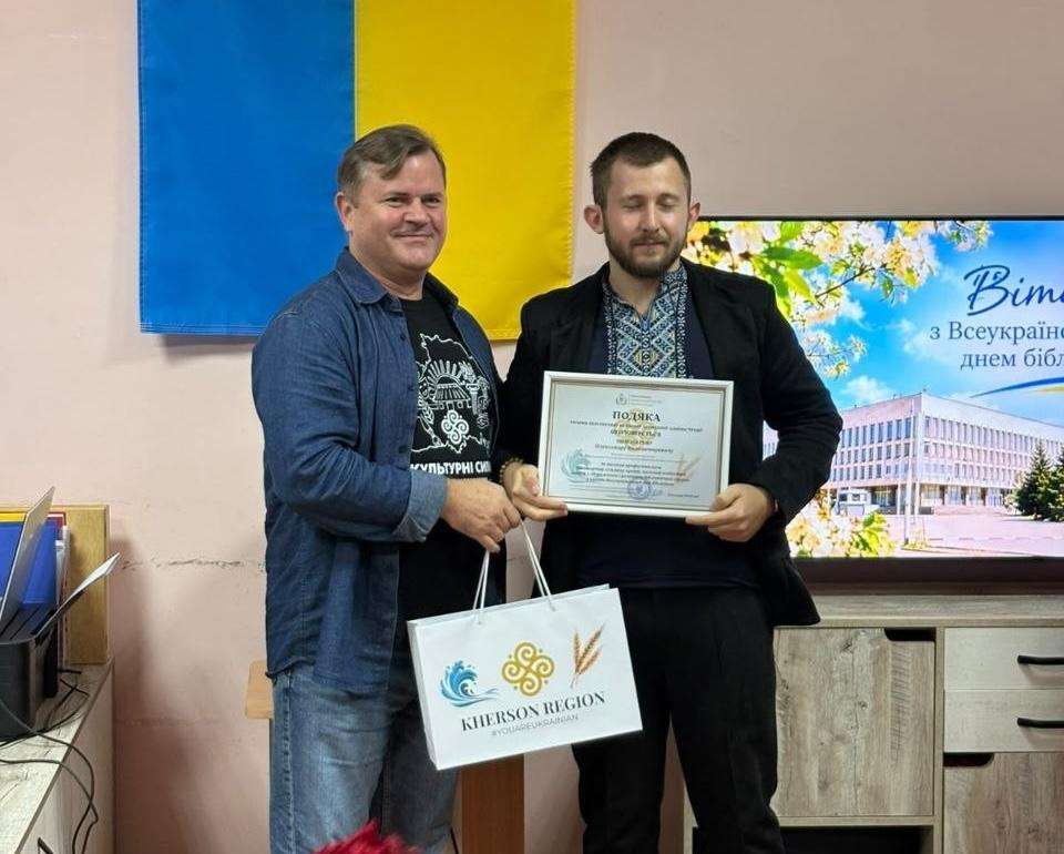 Заступник голови Херсонської ОДА Володимир Клюцевський у синій джинсовій сорочці вручає нагородну подяку та білий фірмовий пакет «KHERSON REGION» представнику культурної сфери у вишиванці під час урочистого заходу «Культурний форпост: вартові українського слова». Подія відбулася у Херсоні напередодні Всеукраїнського дня бібліотек для відзначення працівників Обласної універсальної наукової бібліотеки імені Олеся Гончара. Зустріч проходить у кабінеті під Державним Прапором України поруч із великим екраном, де на тлі квітучих дерев та будівлі ОВА відображається святковий слайд «Вітаємо з Всеукраїнським днем бібліотек», що ілюструє нагородження найкращих фахівців за внесок у розвиток бібліотечної справи.