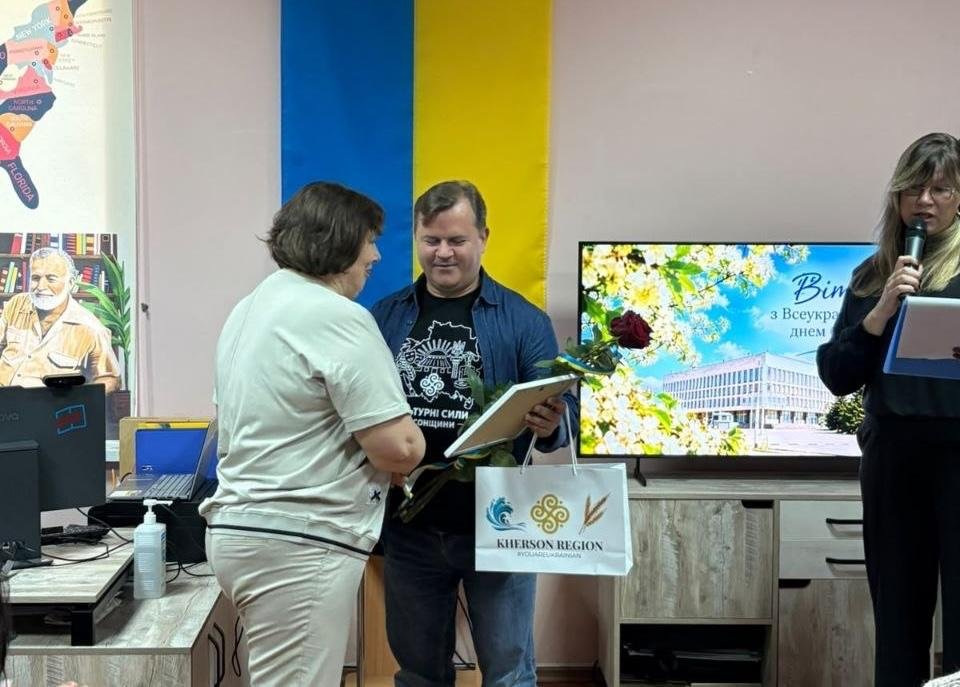 Заступник голови Херсонської ОДА Володимир Клюцевський у синій джинсовій сорочці вручає нагородний диплом у рамці, червону троянду та білий фірмовий пакет «KHERSON REGION» працівниці бібліотечної системи під час урочистого заходу «Культурний форпост: вартові українського слова». Посадовець, на чорній футболці якого нанесено напис «Культурні сили Херсонщини», посміхається під час привітання нагороджуваної жінки у світлому костюмі. Праворуч на передньому плані стоїть ведуча заходу в окулярах, яка зачитує текст із папки у мікрофон на тлі Державного Прапора України та великого екрана зі святковим слайдом «Вітаємо з Всеукраїнським днем бібліотек», що ілюструє відзначення колективу Обласної універсальної наукової бібліотеки імені Олеся Гончара.