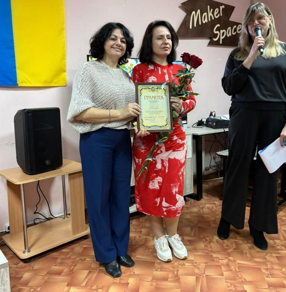Директорка Департаменту реалізації гуманітарної політики Херсонської ОДА Віолетта Царькова у світлій ажурній кофті та синіх штанях щиро посміхається поруч із нагородженою працівницею у яскраво-червоній сукні з білими візерунками під час заходу «Культурний форпост: вартові українського слова». Нагороджувана жінка тримає в руках почесну офіційну грамоту в рамці та букет червоних троянд, перев'язаний патріотичною синьо-жовтою стрічкою. Праворуч на передньому плані стоїть ведуча заходу в окулярах та чорній блузі, яка говорить у мікрофон, тримаючи в іншій руці сценарій. Урочиста церемонія вручення відзнак до Всеукраїнського дня бібліотек проходить у приміщенні на тлі Державного Прапора України, великої аудіоколонки на столику та фірмового дерев'яного покажчика «Maker Space» на стіні, що підкреслює вагомий внесок фахівців Гончарівки у розвиток культури на Херсонщині.