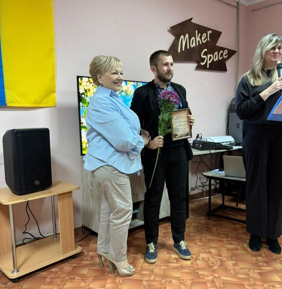 Директорка Херсонської обласної універсальної наукової бібліотеки імені Олеся Гончара Надія Коротун у світло-блакитній сорочці та бежевих штанях щиро посміхається поруч із нагородженим працівником культури у вишиванці та чорному піджаку під час підбиття підсумків конкурсу «Лідери Гончарівки – 2026». Чоловік тримає в руках виборовший диплом у рамці та букет фіолетових хризантем, а праворуч на передньому плані стоїть ведуча заходу в чорній блузі, яка говорить у мікрофон. Урочиста церемонія проходить у приміщенні з фірмовим дерев'яним покажчиком «Maker Space» на стіні, на тлі Державного Прапора України, великої аудіоколонки на підставці та екрана з квітковим вітальним слайдом, що ілюструє проведення заходу «Культурний форпост: вартові українського слова» напередодні Всеукраїнського дня бібліотек.
