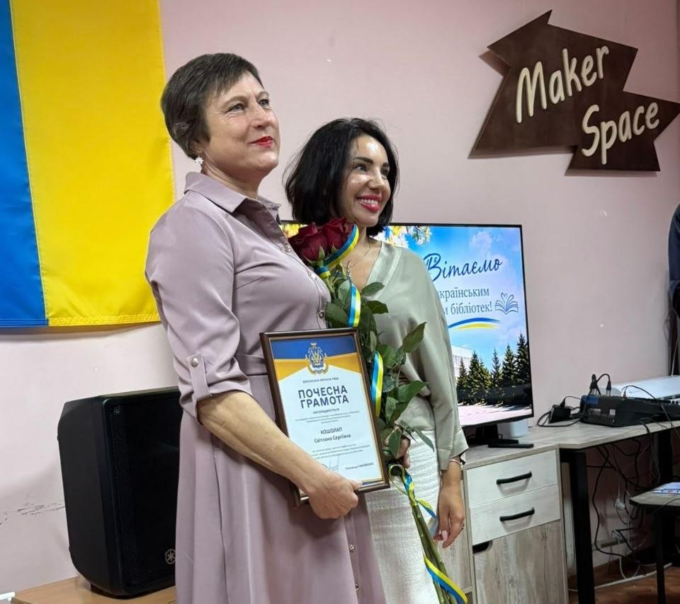Нагороджена працівниця у бузковій сукні тримає в руках «Почесну грамоту» від Херсонської обласної ради та букет червоних троянд із патріотичною стрічкою під час урочистостей до Всеукраїнського дня бібліотек. Поруч із нею посміхається колега у світло-зеленій блузі та білій спідниці під час спільного фотографування у приміщенні хабу «Maker Space». На задньому плані ліворуч висить Державний Прапор України, а праворуч на комоді встановлено великий екран із вітальним святковим слайдом «Вітаємо з Всеукраїнським днем бібліотек», що відображає проведення у Херсоні урочистого заходу «Культурний форпост: вартові українського слова» для підтримки та відзначення колективу Обласної універсальної наукової бібліотеки імені Олеся Гончара.